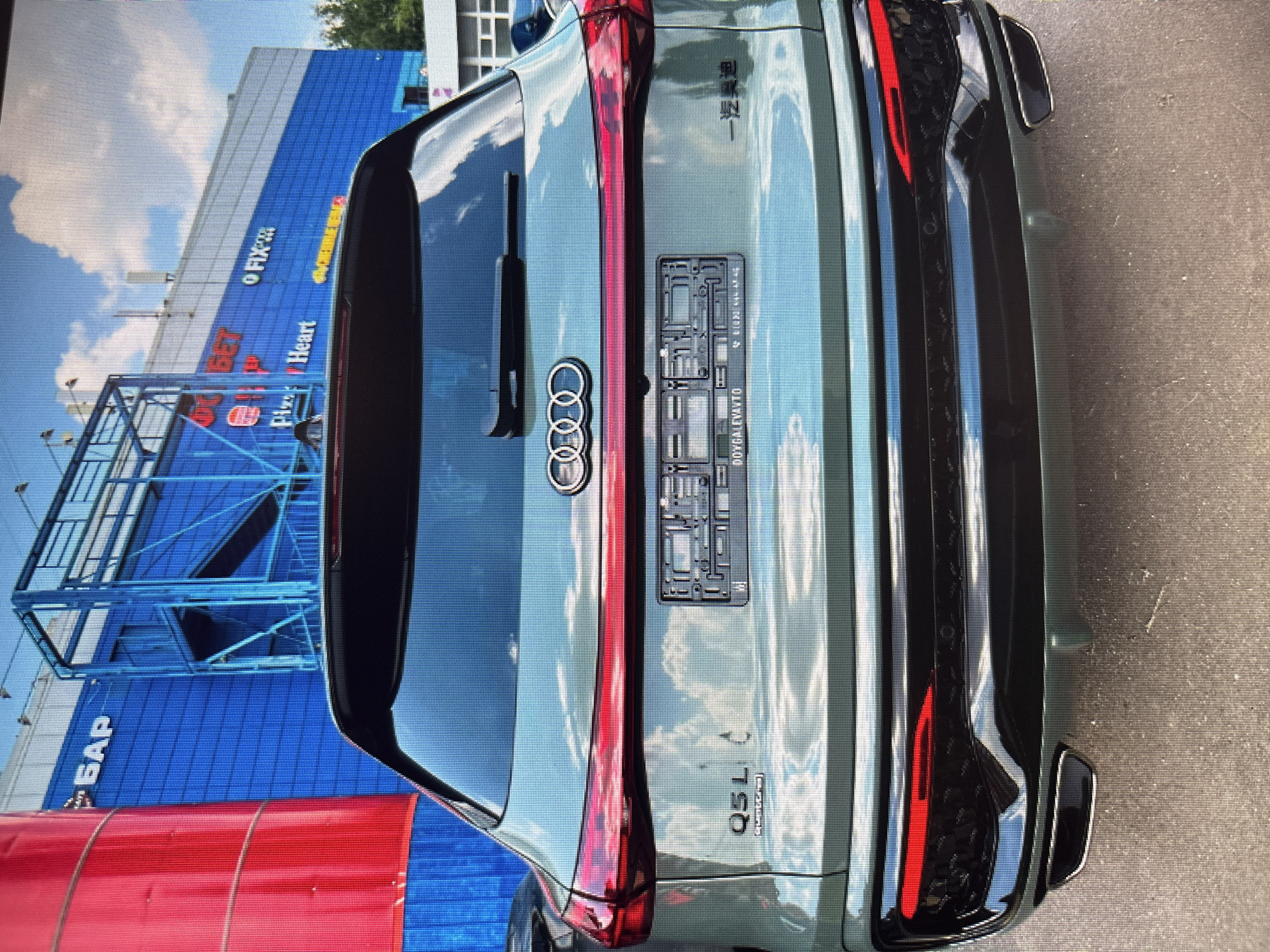 The image shows the rear of a light green Audi Q5L parked on a paved surface outside a building. The building features blue and white facades with red and blue signage. Visible signs include "БАР", "O GET", "Pizz", "Heart", and "FIX". The car has a license plate frame reading "DOYGALEVAVTO" and Chinese characters on the right side. The car's taillights are illuminated red. The sky is visible with clouds, and some trees are present on the right side of the image. The scene appears to be in a commercial area or shopping center.