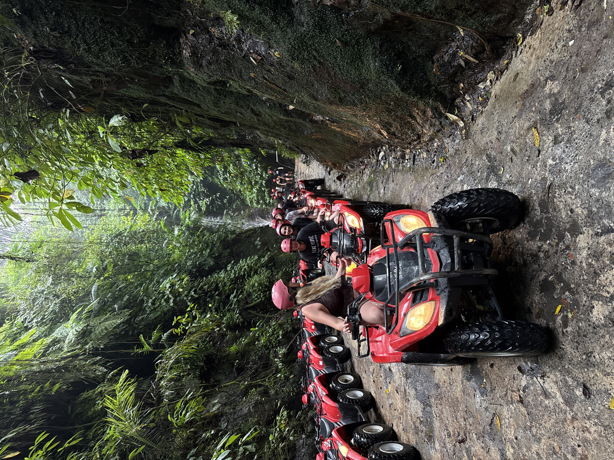 A group of tourists are riding red ATVs through a jungle pathway in Payangan, Indonesia. The path is wet and muddy, with a waterfall visible in the background. The vegetation is lush and dense, with vines hanging from the trees. The ATVs are lined up single file, and the riders are wearing helmets. The image appears to be taken from behind the lead ATV, looking forward at the group and the surrounding scenery. The overall mood of the image is adventurous and exciting.