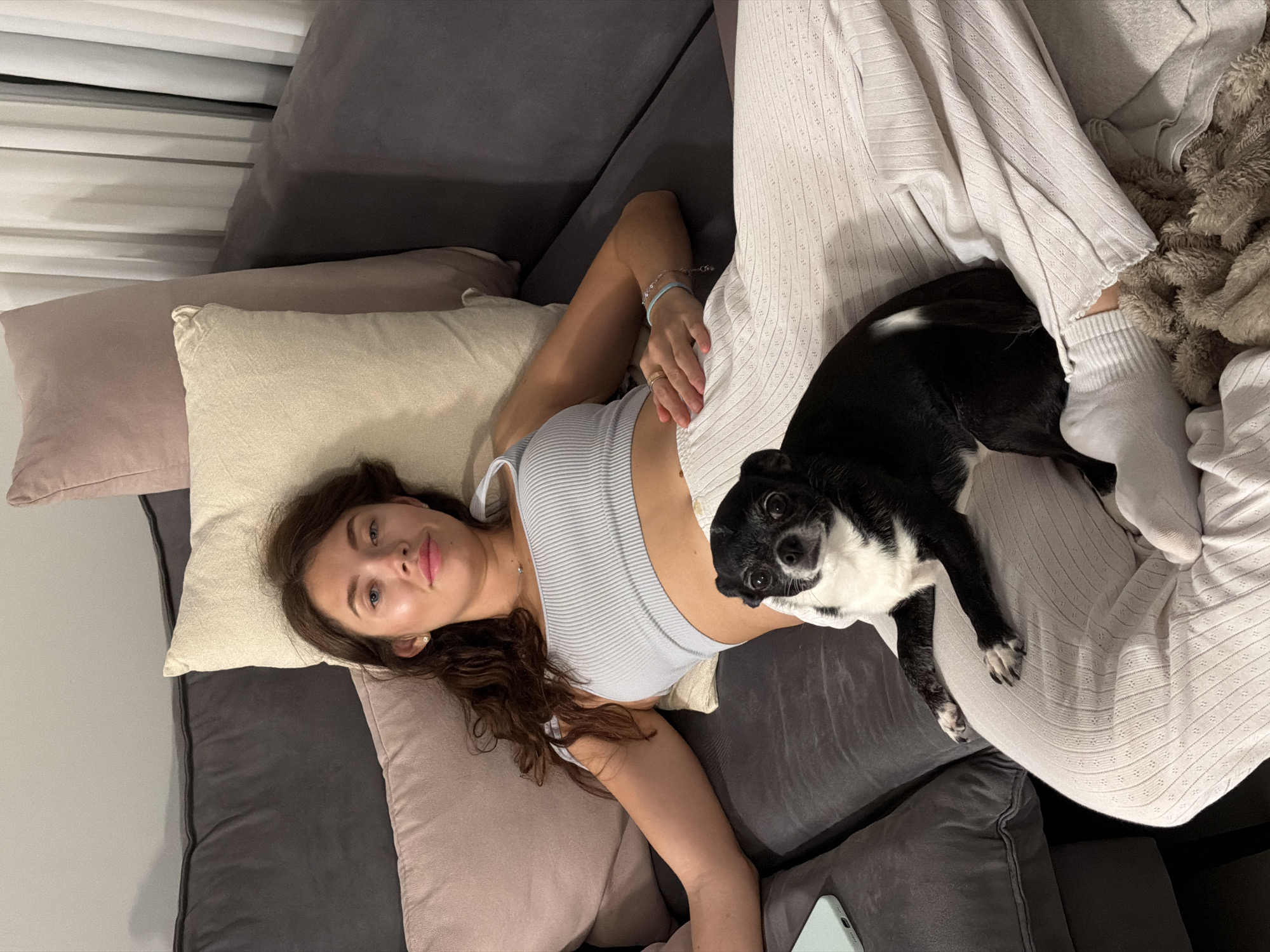 A woman is lying on a gray sectional sofa, surrounded by multiple pillows in shades of beige and pink. She is wearing a light blue ribbed tank top. The woman has brown hair and appears to be gazing at the camera. A small black and white dog is lying on her lap. She is wearing light gray pants and socks. A light blue smartphone is visible near the bottom of the frame. The location in the city of Tel-Aviv, Israel, is unknown from this image.