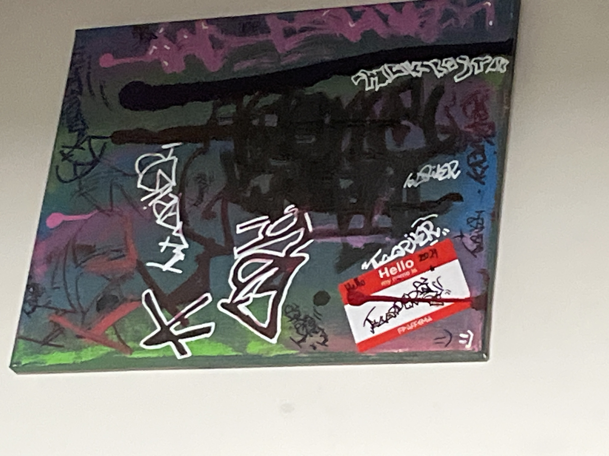 The image shows a square canvas hanging on a white wall. The canvas is painted with a chaotic mix of colors and scribbled text in various styles, including white, black, and other colors. A red and white "Hello" name tag is stuck on the canvas in the lower right corner. It has a year "2021" written on it and other scribbles with a red marker. The painting style seems to be reminiscent of graffiti or street art.
There is no information about the location in the city of Chaikovsky, Russia.