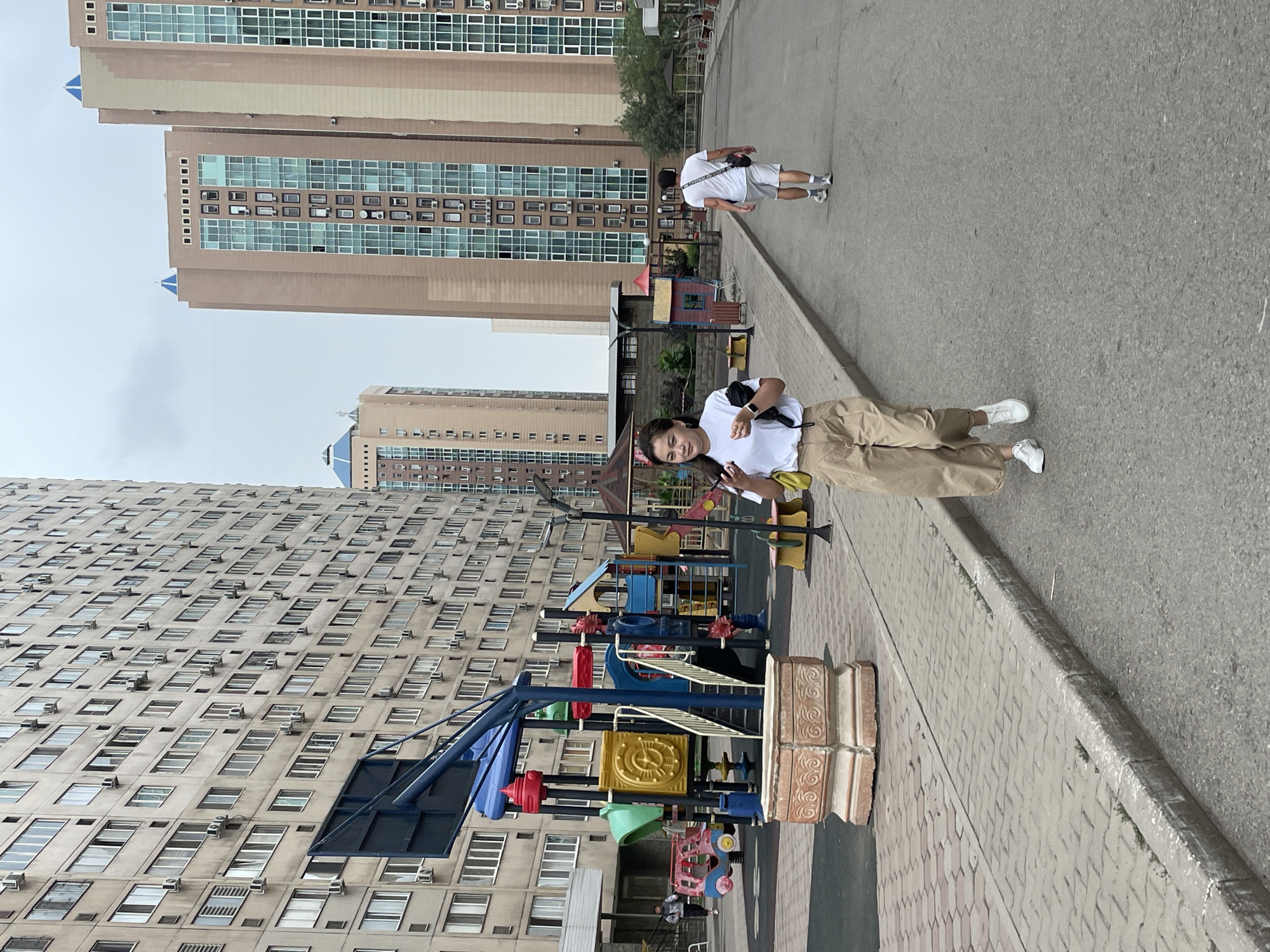 The image depicts an outdoor scene in Almaty, Kazakhstan, featuring residential buildings, a playground, and individuals. In the foreground, a woman is walking towards the camera, holding a phone and wearing a white t-shirt and beige pants. Behind her, a man in a white t-shirt and shorts is walking away from the camera.
The environment includes a paved road and a sidewalk. To the left, a colorful playground structure is visible with slides and climbing elements. Behind the playground, a large, multi-story residential building with numerous windows and air conditioning units dominates the background on the left. On the right side of the image, several modern, high-rise apartment buildings with tan and green facades are present. The sky is overcast.