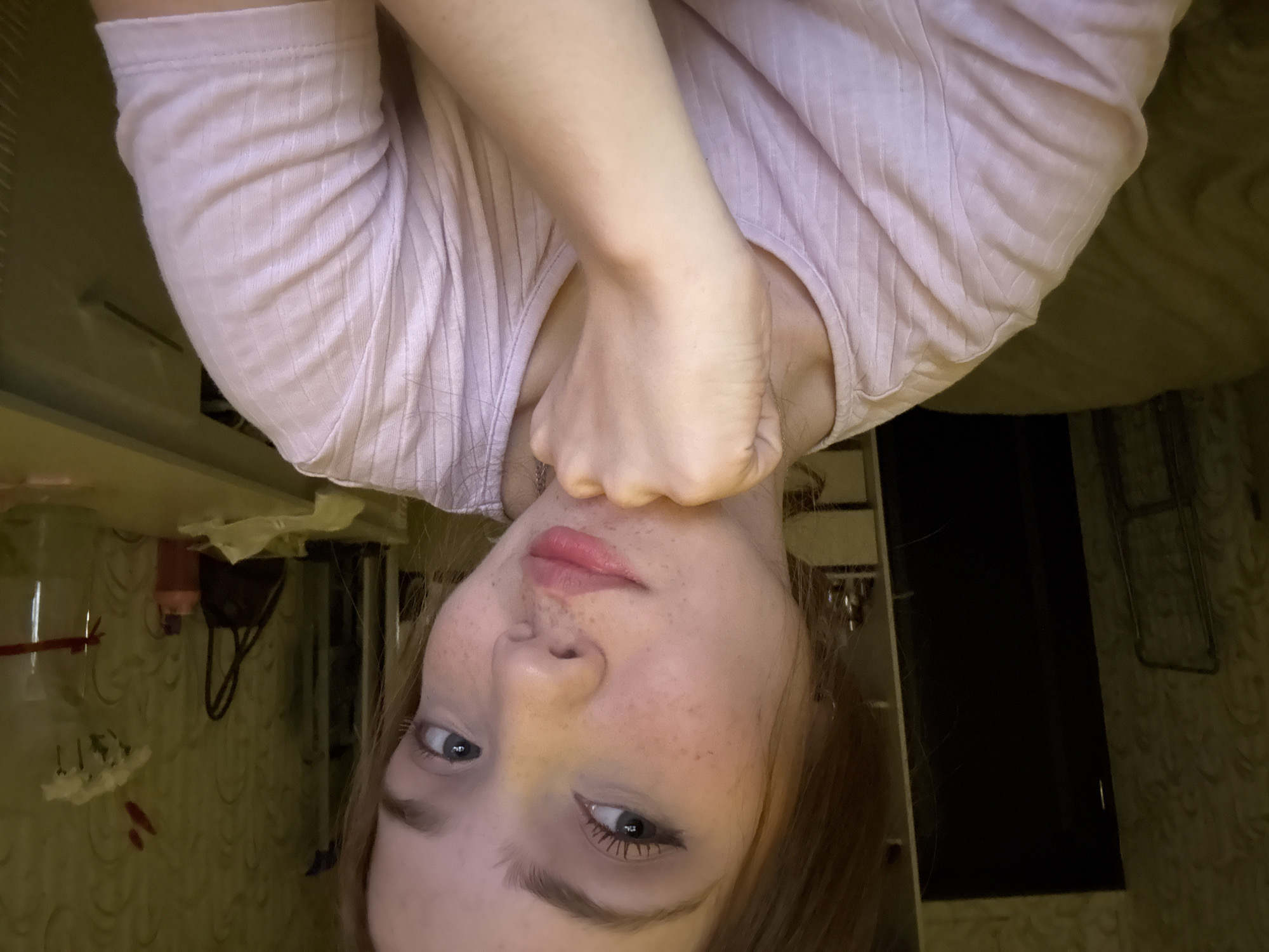 A young adult female, appearing to be in her late teens or early twenties, is positioned centrally in a medium-shot portrait within an indoor setting. She has light skin with numerous freckles across her nose and cheeks, light-colored eyes with visible winged eyeliner and mascara, and light brown or auburn hair that falls to her shoulders. Her lips are lightly colored with pink lipstick, slightly parted, and she maintains a direct gaze towards the viewer. Her right hand is clenched into a fist and rests under her chin, while her left arm is partially visible on the left side of the frame. She is wearing a light pink, ribbed, short-sleeved top with a rounded neckline.
The background depicts a room with patterned wallpaper, possibly a floral or damask design, in a muted olive-green or beige tone. To the left, a tall, dark, rectangular object, likely a door or wardrobe, is visible, with a faint vertical light reflection on its left edge. Below it, a metallic, foldable object, possibly a drying rack, rests against the wall. Behind the subject's left shoulder, a light-colored shelving unit or set of drawers holds small, reflective objects. To the right of the subject, a light-colored table or dresser surface extends across the frame. On this surface, from left to right, are a dark handbag with a curved handle, a pink/orange plastic bottle, a crumpled white tissue, and a tall, clear glass container, possibly a vase, with a red ribbon tied around its neck. Partially obscured by the clear container is a small arrangement of white flowers with red accents. The lighting is artificial and somewhat dim, casting subtle shadows. Below the subject's right arm, a textured fabric, possibly bedding, in a light beige or olive color is visible.