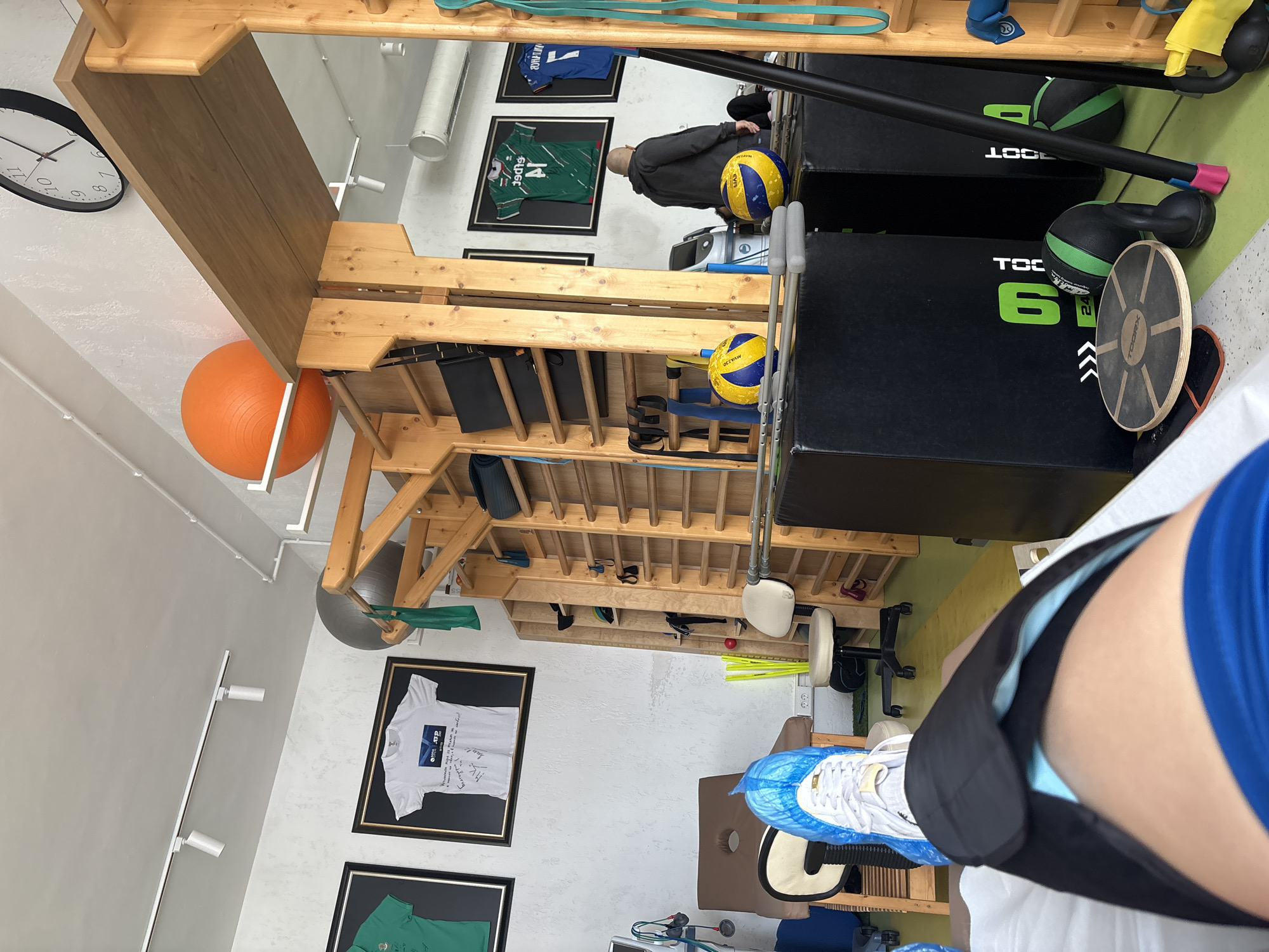 The image displays the interior of what appears to be a physiotherapy clinic, gym, or rehabilitation center, filled with various exercise and therapy equipment. The photo is rotated 90 degrees clockwise, with the floor running along the right side of the frame and the ceiling along the left.
In the immediate foreground, a person's lower right leg and foot are visible. The person wears a blue short-sleeved shirt and a black leg brace or compression sleeve around their thigh and knee. Their foot, wearing a white sneaker covered by a blue disposable shoe cover, rests on a brown and white therapy table or piece of equipment.
The midground and background showcase a range of items. A prominent wooden shelving unit or wall bars structure stretches across the center of the room. On what would be the floor (right side of the image), two black plyo boxes are stacked, one bearing the logo "TOORX" and the number "6". Adjacent to these are two black medicine balls with green highlights, a long black bar, and a wooden balance board. A pair of crutches stands upright. Two yellow and blue volleyballs, branded "MIKASA", are visible, one on a shelf and another on the floor. An orange exercise ball is suspended near the ceiling (left side of the image), and a grey exercise ball is partially visible under the wooden structure.
In the background, a person wearing a dark hoodie stands with their back to the camera. The white walls are decorated with several framed sports jerseys and t-shirts; these include a green jersey with "14" and "SPORTS" printed on it, a blue jersey, and a white t-shirt with "ATP" text. A black and white wall clock, indicating approximately 1:30, is mounted on the wall. Overhead, track lighting fixtures are visible. Green, yellow, and blue resistance bands are scattered throughout the space, suggesting a focus on physical rehabilitation and training.