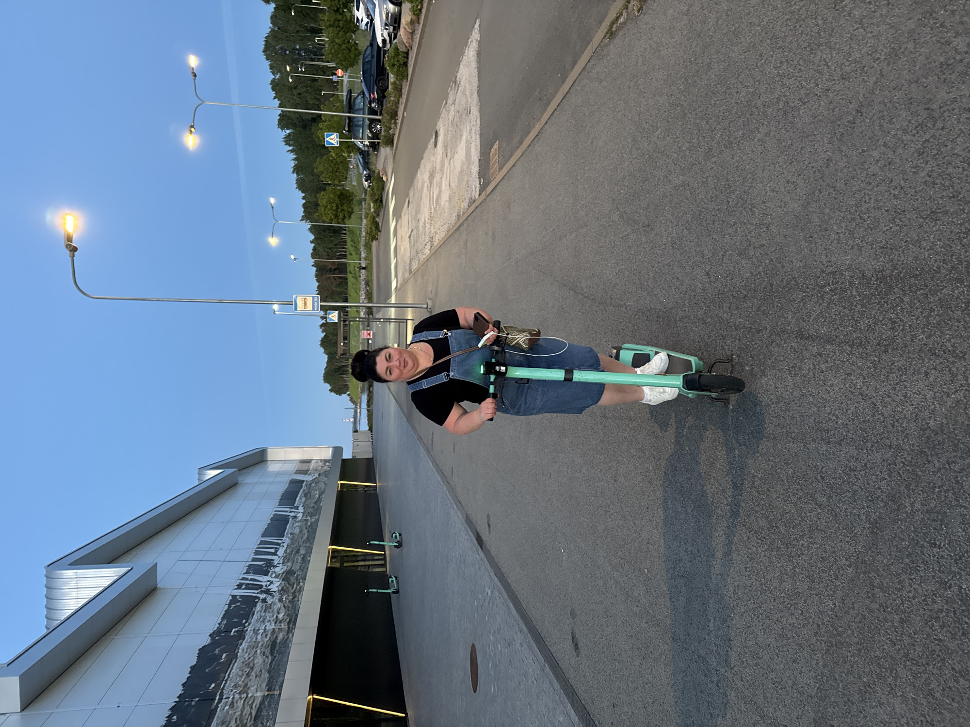 A woman with dark hair styled in a bun rides a green electric scooter down an asphalt path in Tallinn, Estonia. She wears a black t-shirt and denim overalls. Her right hand is on the scooter's handlebars, and she holds a phone in her left hand. The scooter casts a shadow on the ground to her left.
The path is bordered by a large, modern building with a metallic facade on the left and a parking lot with cars and trees on the right. Several other green electric scooters are parked near the building. Streetlights illuminate the scene, and the sky is a clear, deep blue. The overall lighting suggests it is dusk or early evening.