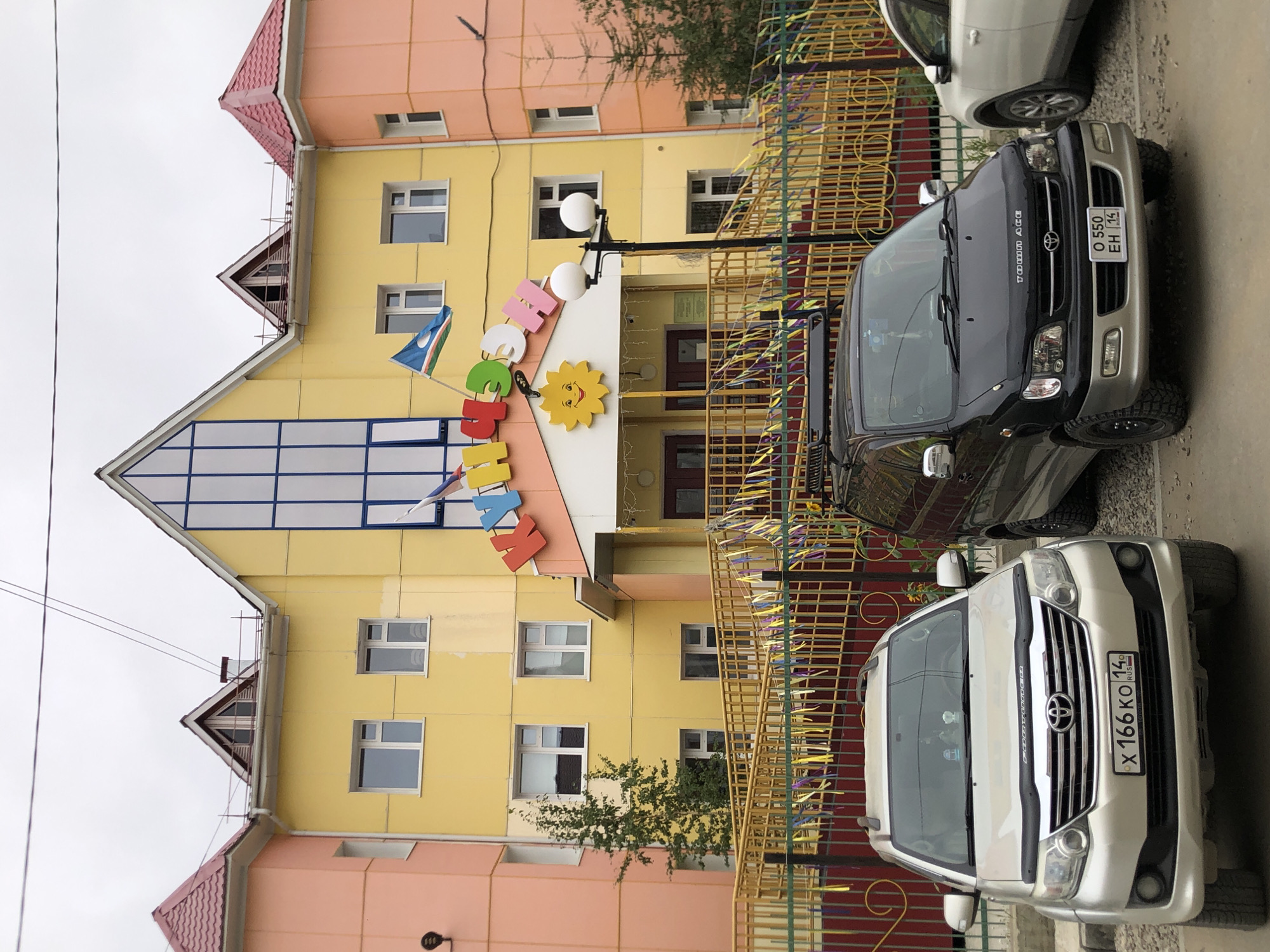 The image shows a bright yellow and pink building with a red roof, resembling a kindergarten or daycare center. Colorful letters spelling out "КЛУМБА" (Klumba), meaning "flowerbed" in Russian, are prominently displayed above the entrance. A large yellow sun decoration is also visible. Two cars are parked in front of the building: a silver Toyota Fortuner on the left and a black Toyota Town Ace on the right. Both vehicles have license plates that appear to be from Russia, specifically showing "RUS". The building is located on a street with a fence decorated with colorful ribbons. The environment suggests an overcast day in Yakutsk, Russia, a city known for its extreme cold. The scene captures a typical urban setting with a focus on the entrance of a children's facility and the vehicles present.