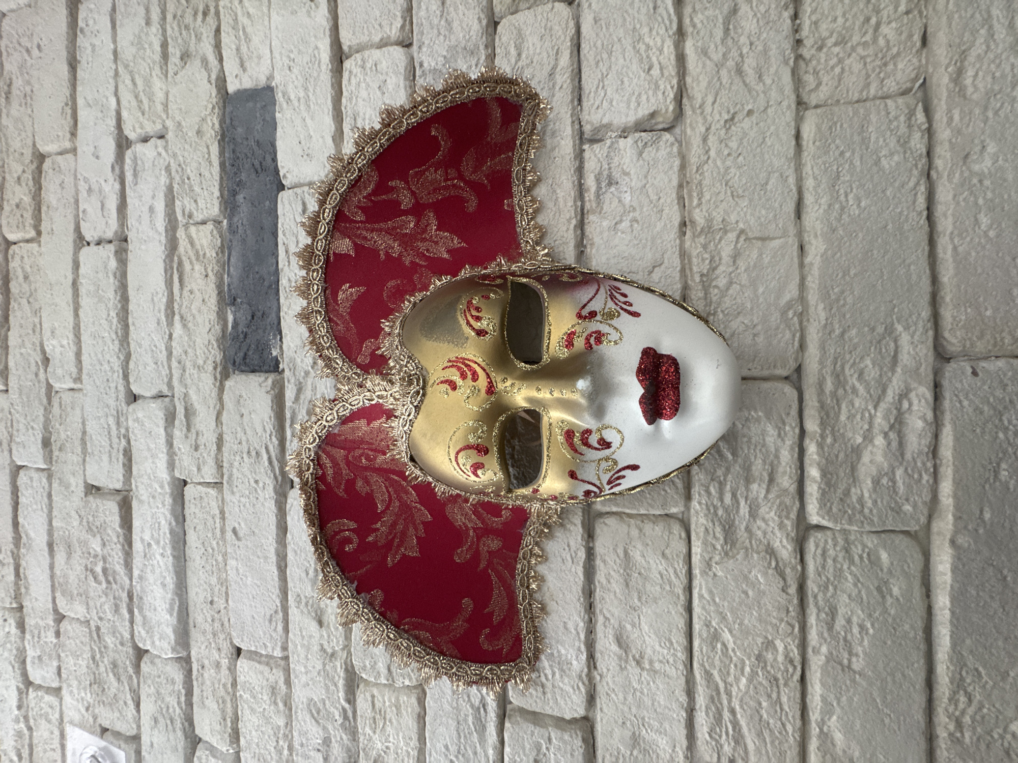 A close-up, eye-level, medium shot captures a decorative Venetian mask hanging on a white brick wall. The mask is positioned centrally, dominating the frame. The wall features a roughly textured, white brick pattern with visible mortar lines. A single dark gray brick is noticeable in the upper middle section of the wall.
The mask itself is ornate, with a white porcelain face. The upper portion and eye sockets are covered in shimmering gold, adorned with intricate red scrollwork and glittering red accents. The cheeks feature delicate red designs that extend towards the eyes. The lips are a prominent feature, painted in a sparkly, deep red. Attached to the sides of the mask are large, fan-shaped decorative elements made of red fabric with a gold damask pattern. These fabric pieces are edged with a gold, intricate lace trim. The overall impression of the mask is one of festivity and theatricality.
The lighting appears to be even and diffused, suggesting indoor lighting or an overcast day, casting no harsh shadows. The background of the brick wall is in sharp focus, as is the mask. No people are present in the image, and no actions are being performed. The setting is static and appears to be a wall in a residential or commercial space, possibly a home in Agios Tychona Community, Cyprus.