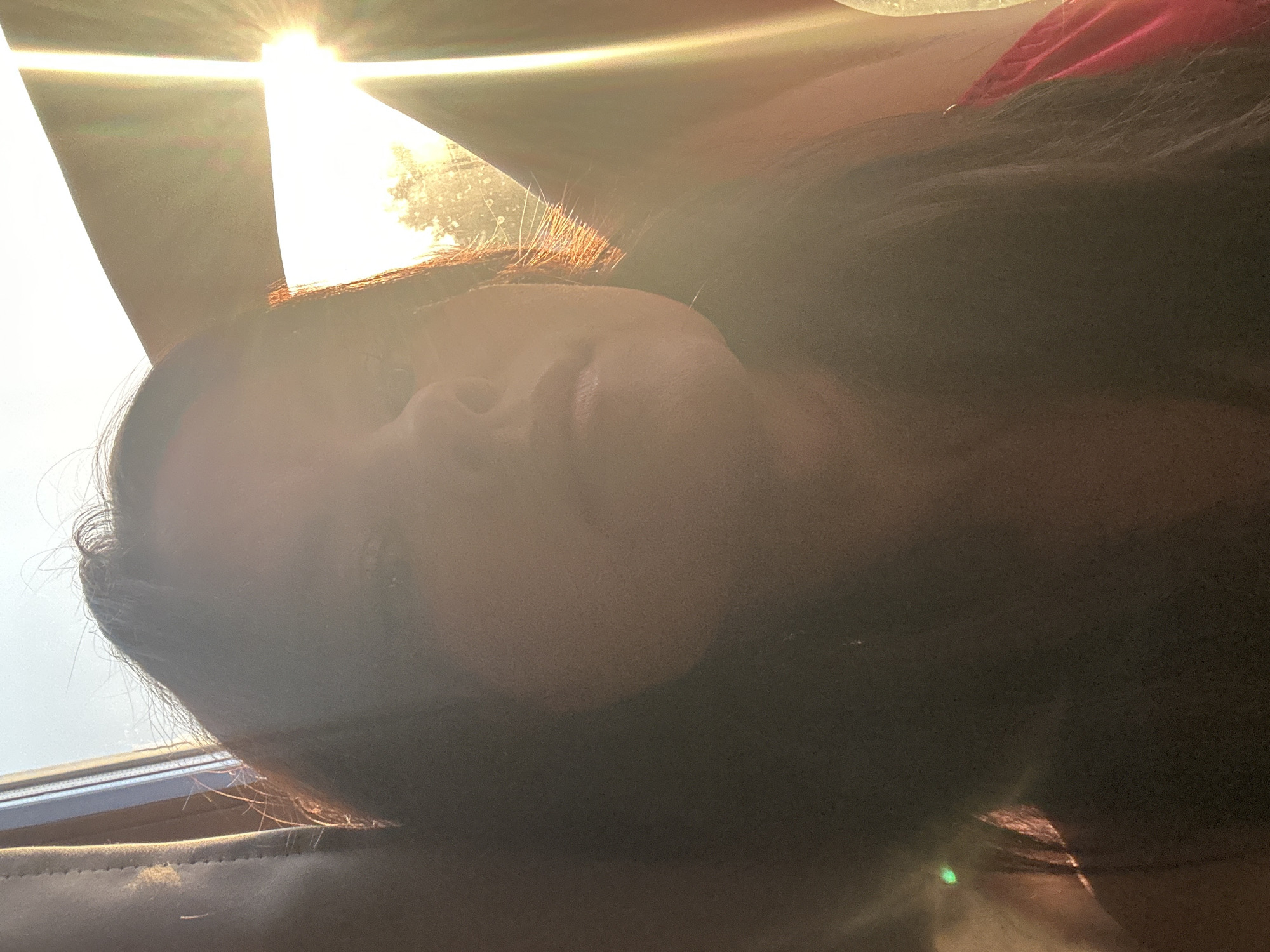 The image presents a close-up portrait of a person, likely female, captured from a low angle, suggesting they are resting or reclining. The subject's face is partially obscured by strong, direct sunlight streaming from the upper left, creating a prominent lens flare and overexposed areas. This intense backlighting illuminates portions of the person's dark hair, causing some strands to glow with a reddish-brown hue. The rest of the face and hair are cast in shadow, creating a high-contrast effect.
A hint of a red garment is visible in the upper right portion of the frame. The setting appears to be the interior of a vehicle, possibly a car, indicated by what resembles seat upholstery or a headrest in the bottom left and a window frame structure in the upper left. The bright sunlight strongly suggests it is daytime, likely a clear and sunny day. The person's posture implies a state of relaxation or calm. No specific activities are depicted beyond this relaxed state, and no text is visible within the image. This scene takes place in Chaikovsky, Russia.