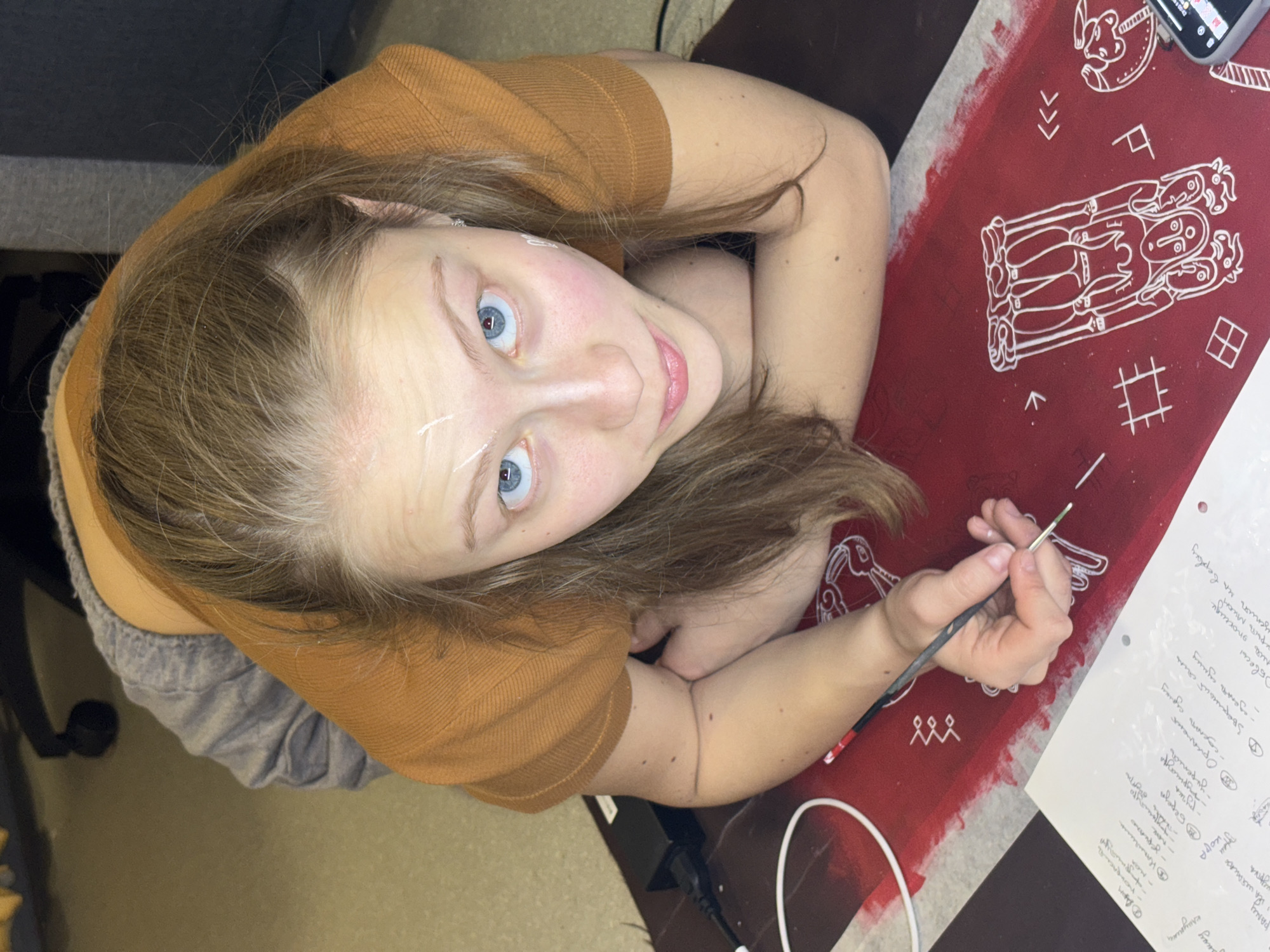 A woman with fair skin, light brown hair, and striking blue eyes is painting intricate white designs onto a red cloth. She's wearing a tan-colored, long-sleeved shirt and gray shorts and appears to be working at a desk or table, with a piece of paper containing handwritten notes beside her. The notes are in Russian. The cloth design includes stylized figures and geometric patterns. The scene may be located in Perm, Russia, based on the potential context of the Russian text.