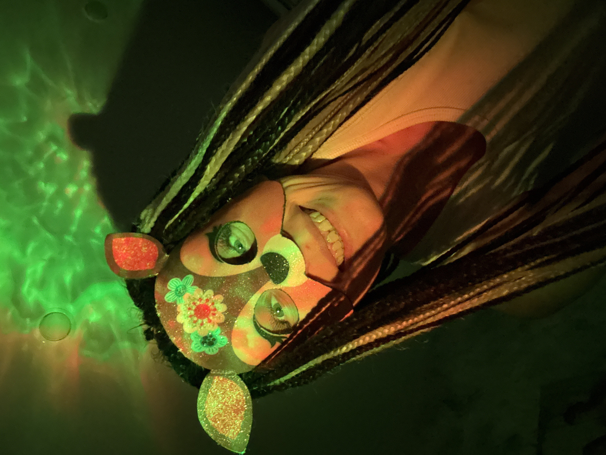 A headshot of a person is shown, captured at an unusual, tilted angle, giving the impression of being inverted. The subject has long, distinctive braided hair, featuring a mix of dark and lighter strands, possibly synthetic dreadlocks. Their face is elaborately decorated with a glittery, mask-like makeup design, incorporating ear-like shapes, round eye cutouts, and colorful floral patterns (red and green) on the cheeks. The person's eyes are visible and look directly towards the viewer, and they are smiling broadly, showing a full set of teeth, indicating happiness or amusement.
The scene is characterized by dramatic lighting. A strong green light source illuminates the left side of the frame, creating wavy, dappled patterns on a surface and casting a vibrant green hue across the background and parts of the subject. The person's skin tone appears warm, contrasting with the cool green light. Shadows are deep and pronounced, enhancing the dramatic atmosphere. The setting appears to be indoors, likely a venue such as a party, club, or performance space, given the artificial and colorful lighting. There are no visible texts or specific location cues within the image itself. The overall impression is one of festivity and joyful celebration, taking place in Rusanovo, Russia.