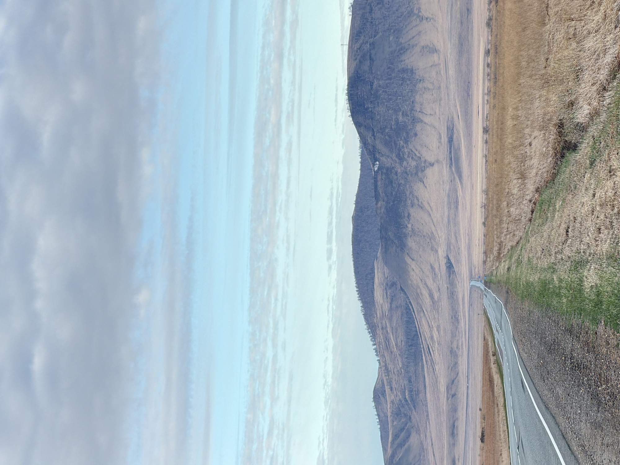A straight, asphalt road with a white line marking the shoulder runs between a tan, grassy field on the right and a barren, sloping hillside on the left. The sky above is a mix of light blue and white, with clouds streaked horizontally across the view. No people or other animate objects are visible. The environment appears rural and natural, devoid of any urban or constructed elements specific to Aeshka, Russia.