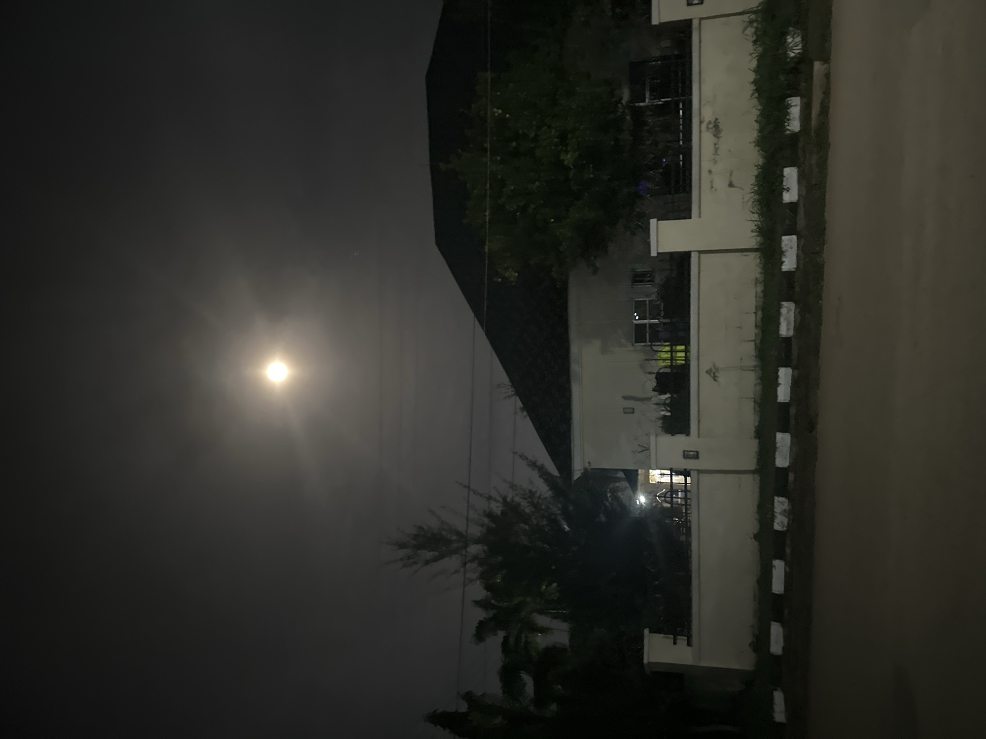 The image shows a residential building in Abuja, Nigeria, at night under a hazy full moon. The building is a single-story structure with a dark tiled roof and cream-colored walls. It has several windows, some of which are illuminated. A white perimeter wall surrounds the property, with black and white striped bollards lining the sidewalk in front of it. Trees, including palm trees and a tall conifer, are visible on the property. The street in front of the house is paved. The overall scene suggests a quiet, residential area at night.
