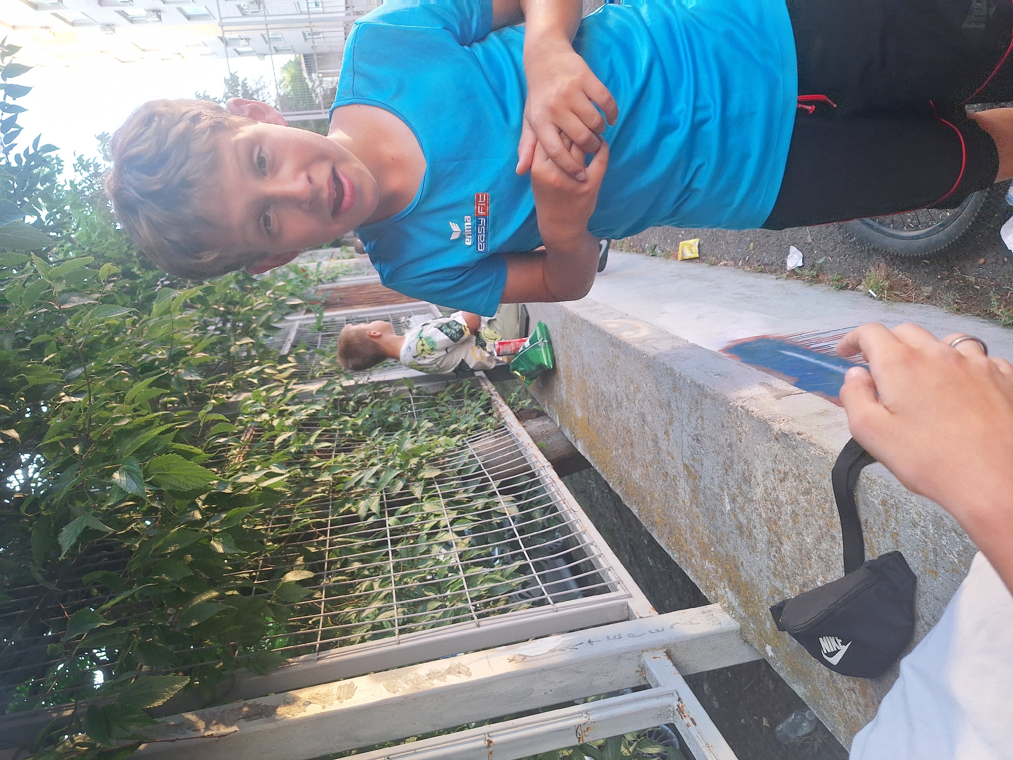 The image depicts a young boy with light brown hair and fair skin, wearing a bright blue t-shirt with a red and white logo on the left chest, and black shorts with red trim. He is standing outdoors in what appears to be an urban environment. In the background, another young boy is visible, facing away from the camera. The setting includes a metal fence covered in green foliage, a concrete ledge, and a building in the distance. On the concrete ledge, there is a black fanny pack with a white Nike logo. The ground is dirt with some scattered debris. The overall lighting suggests it is daytime, possibly late afternoon due to the shadows. This scene is consistent with the area near the Yambol Central Bus Station in Yambol, Bulgaria, which features similar fencing, concrete structures, and urban vegetation.