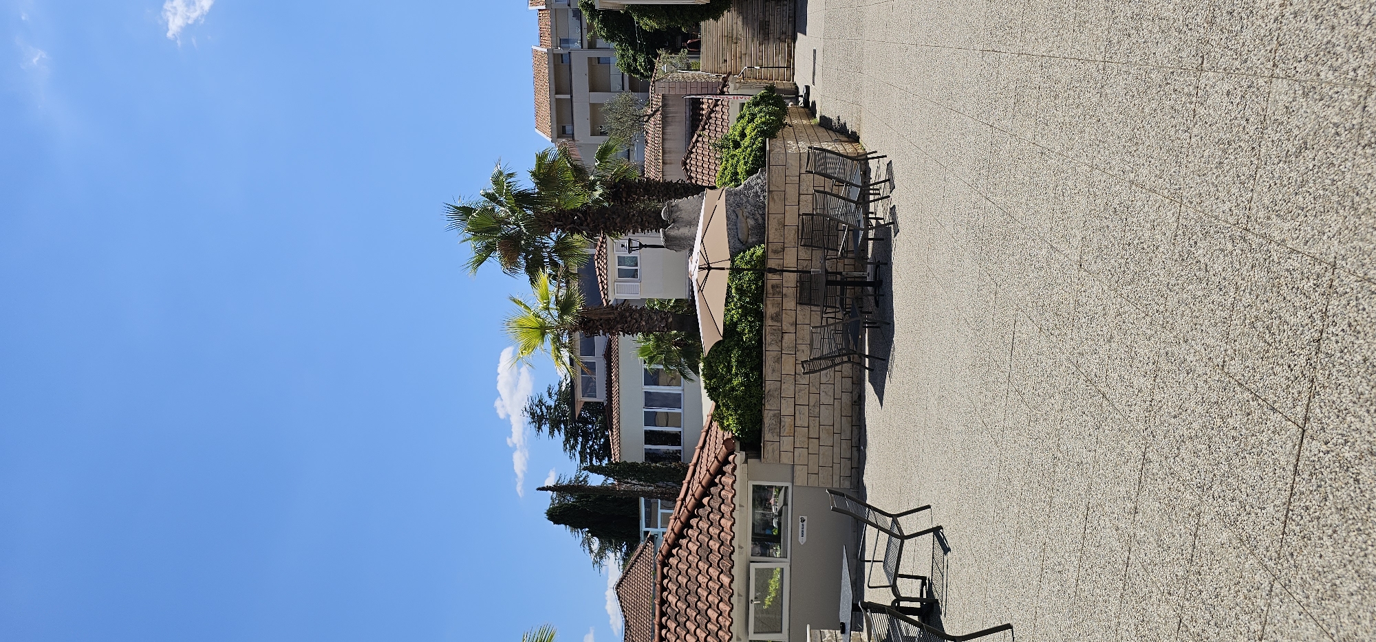 This is a full-shot, exterior, eye-level photograph taken in the city of Grad Supetar, Croatia. The scene depicts a bright, sunny day with a clear blue sky. In the foreground, a textured, light-colored paved area dominates the lower half of the image. Several dark metal tables and chairs are arranged on this pavement, some grouped around a large, light-colored umbrella.
Behind the seating area, a low wall constructed of rectangular stone blocks separates the paved area from a lush green garden. Two palm trees with vibrant green fronds rise above the garden. In the background, several buildings with terracotta tiled roofs and light-colored facades are visible. The buildings appear to be part of a resort or hotel complex, with balconies and large windows. To the right, a stone staircase leads upwards. A single person is visible walking up the stairs in the far right background. The overall impression is of an outdoor seating area within a Mediterranean resort.