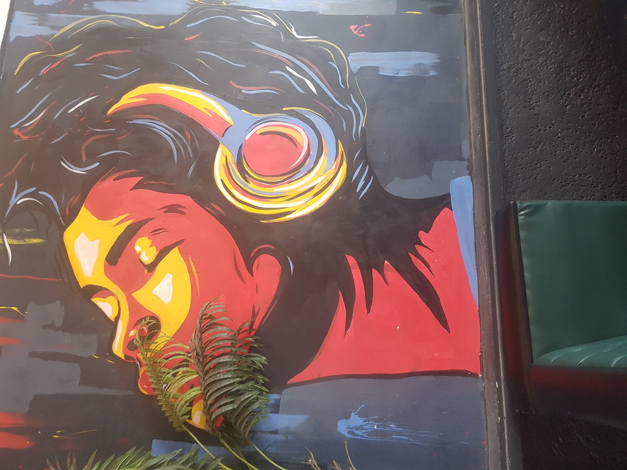 This image depicts a colorful mural painted on a dark wall, featuring a stylized portrait of a person wearing headphones. The face is rendered in bold strokes of red, yellow, and white, with blue accents. Lush green foliage partially obscures the left side of the mural. The lower portion of the image shows a dark textured wall below the mural, and a hint of a green upholstered surface, possibly a seat or bench. The mural's vibrant colors and modern design suggest it is a piece of urban art. The specific location in Howayi, Nigeria cannot be determined from the image alone.