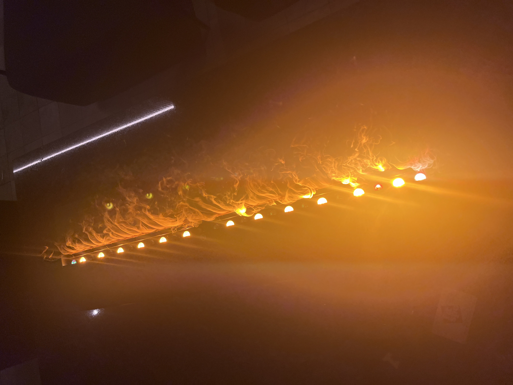 The image displays an atmospheric indoor scene, likely a performance venue, characterized by dramatic artificial lighting and smoke effects. A prominent feature is a horizontal row of at least fifteen bright, semi-circular yellow-orange lights, possibly stage fixtures, casting a warm glow. Above these lights, a textured, wavy material, likely fabric or smoke, is illuminated from behind, creating organic, flame-like patterns that extend across the frame. Notably, several distinct glowing green circular shapes, resembling eyes, are visible within these patterns on the left side.
In the upper-left quadrant, a contrasting linear white light strip, possibly an LED or neon fixture, runs diagonally against a dark background. The lower portion of the image is deeply shadowed, suggesting a floor or unlit area. On the far right, a very faint, rectangular shape can be discerned, possibly a poster or sign, but any visible text is too indistinct to read.
The scene conveys the vibrant ambiance of a nighttime event, such as a concert, club, or live performance, with the intense artificial illumination and smoky effects contributing to an energetic and immersive atmosphere in Sofia, Bulgaria.