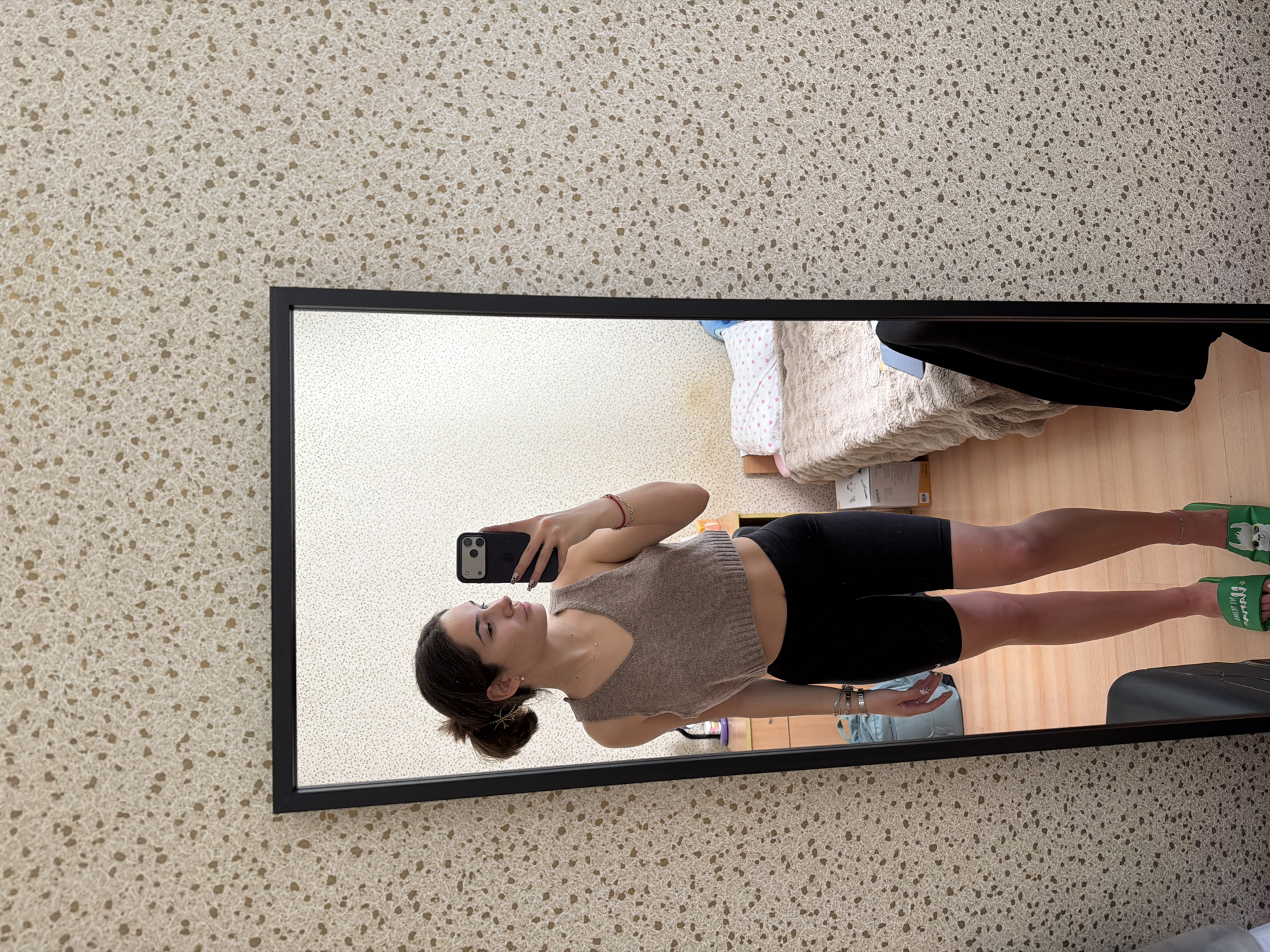 A young woman is taking a selfie in a full-length mirror. She is wearing a tan tank top and black shorts with green slippers. The mirror is reflecting a bedroom scene with a bed, a dresser, and a chair. The wall behind the mirror has a speckled beige pattern. The photograph may have been taken in Sofia, Bulgaria, but there is nothing in the picture to confirm the specific location.