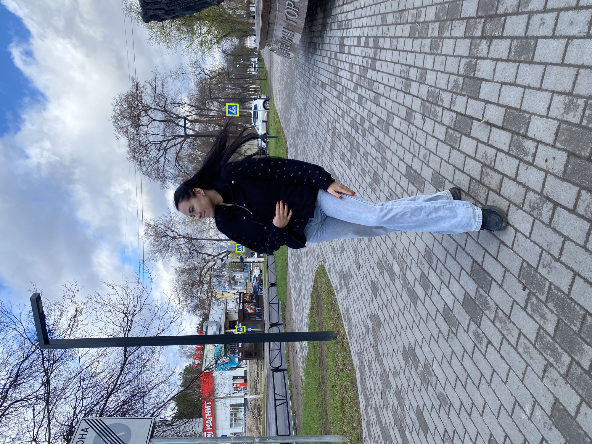 The image, correctly oriented, displays a young woman standing on a paved pedestrian path in an outdoor urban or park-like setting in Chaikovsky, Russia. She has long, dark hair that appears to be blowing to her right, suggesting a breeze. She is wearing a black jacket with subtle patterns, light blue jeans, and dark shoes, with red nail polish visible on her hands, which are placed near her stomach. Her gaze is directed slightly downwards and to her left.
The path is constructed from alternating light and dark grey rectangular pavers. To the woman's right, a grassy strip separates the path from a road, where a white car and a yellow pedestrian crossing sign are visible. In the background, various buildings line the street, including one with a red facade featuring white text that clearly reads "МАГНИТ" (Magnit), indicating a supermarket.
To the woman's left, another grassy area borders a large, dark monument or plaque. Partially visible text on this structure reads "...ЧАЙКОВСКИЙ ГОР...", likely part of "ЧАЙКОВСКИЙ ГОРОД" (Chaikovsky City/Town). The scene is framed by numerous bare trees with intricate branches against an overcast sky that shows patches of blue and white clouds, suggesting daytime, possibly during the transitional seasons of early spring or late autumn. In the immediate foreground on the left, a dark metal post supports a partially visible white road sign displaying "ОНА" and a diagonal line pattern, possibly indicating the end of a zone or specific regulation. The atmosphere is that of a typical, albeit breezy, day in a town.