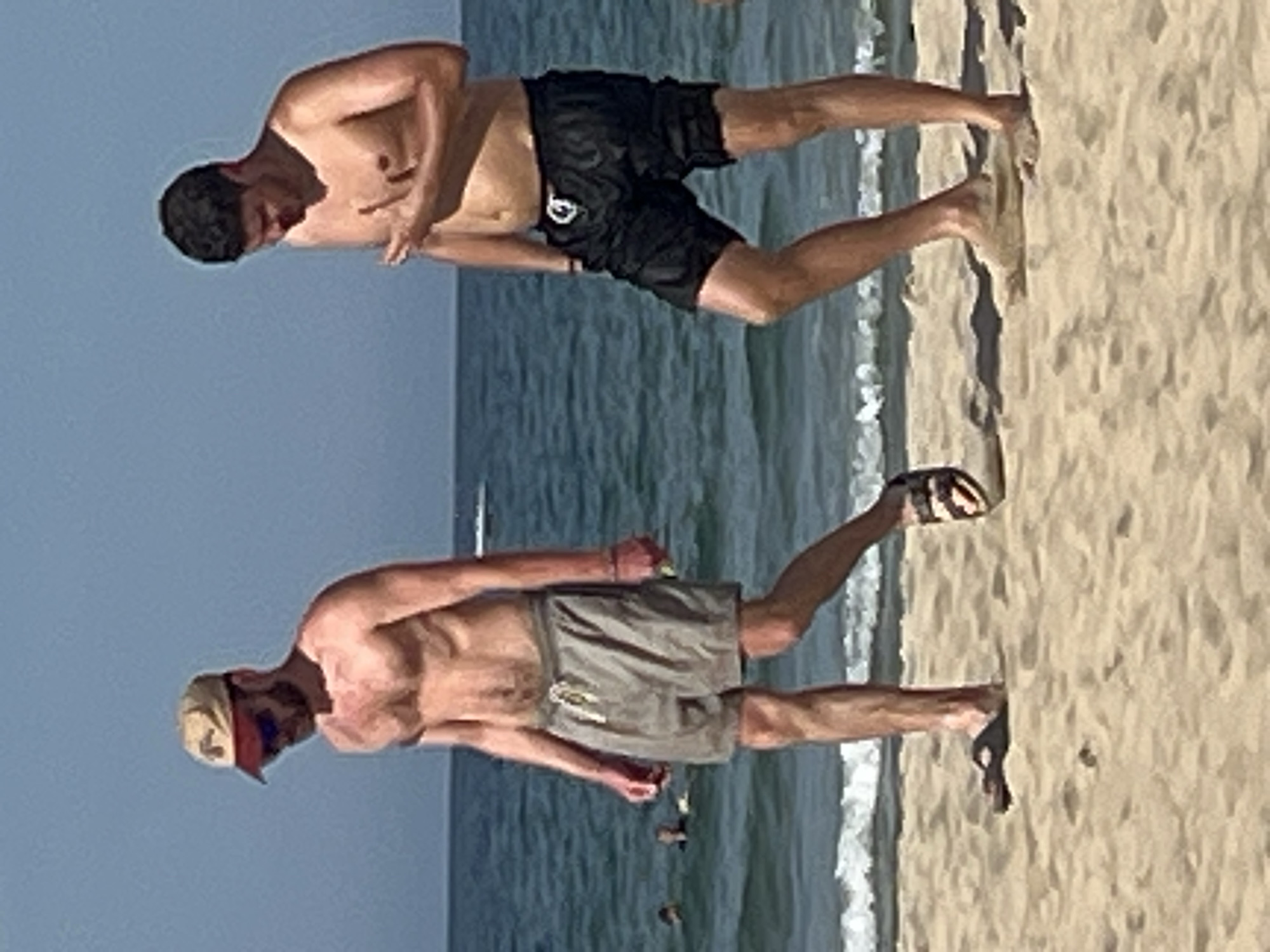 Two men are walking on a sandy beach in Da Nang, Vietnam. The man on the left is wearing a tan baseball cap, sunglasses, and grey swim trunks. He has a beard and appears to be looking down. The man on the right is wearing black swim trunks and has his right hand raised to his chest. Both men are shirtless, exposing their torsos. The ocean is visible in the background with small waves breaking on the shore. The sky is clear and blue. There are a few other people in the water in the distance.