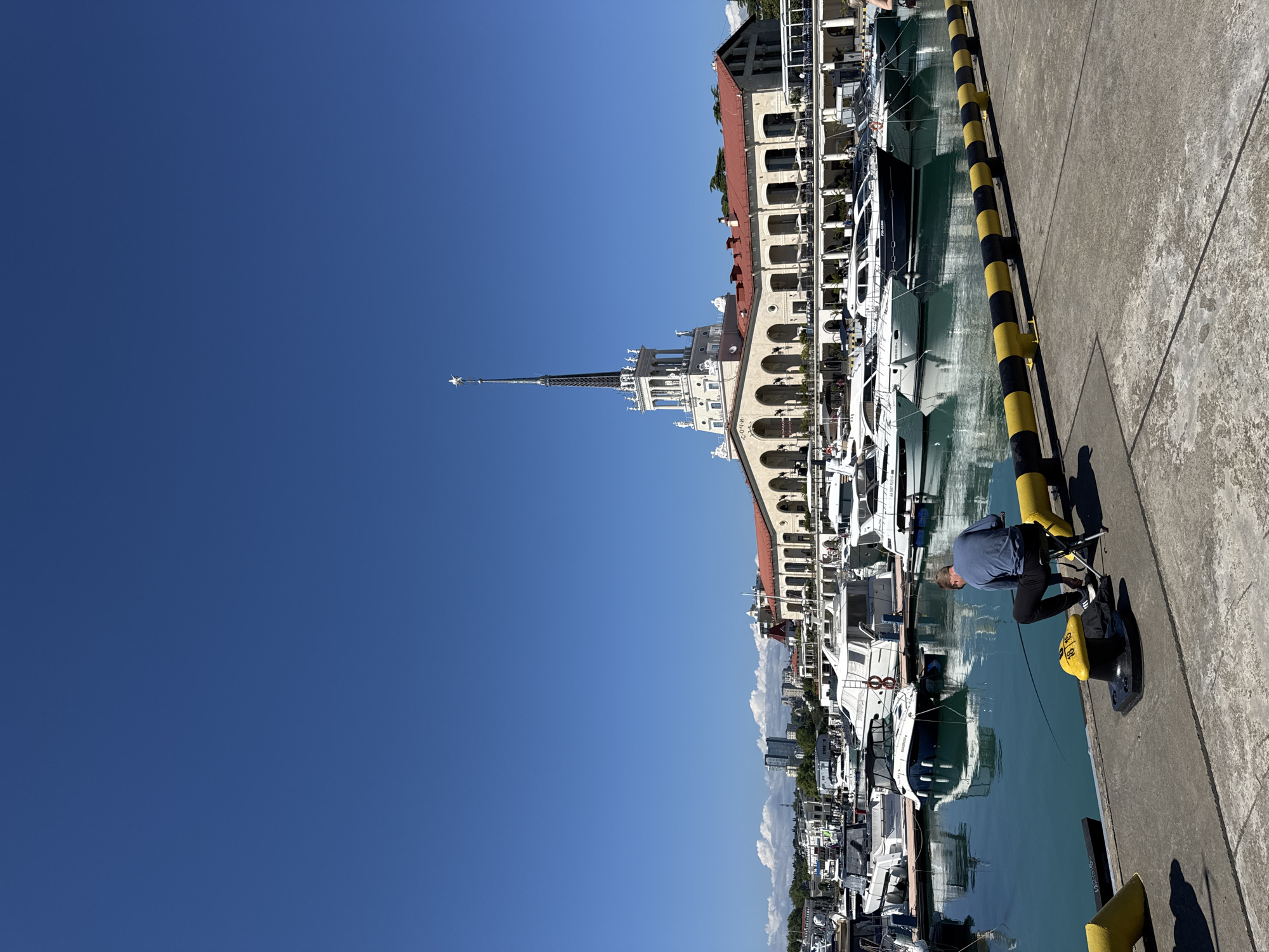 A sunlit, wide-angle view captures a lively marina or port on a clear day. The image is dominated by a vast, cloudless blue sky on the left, covering approximately half of the frame. On the right, an ornate, multi-story building with classical architecture, including arches, columns, and a reddish-brown tiled roof, stretches along the waterfront. A prominent, white spired tower rises from this building, extending into the clear sky. The word "СОЧИ" is visible on the facade of this main building.
Below the buildings, the calm, turquoise-green water of the marina reflects portions of the architecture and the docked boats. A long line of white yachts and other smaller boats are moored neatly along the concrete pier. The pier itself, made of concrete, runs along the bottom and right side of the image. Distinctive black and yellow striped cylindrical bollards are spaced along the edge of the pier.
In the lower right corner, a man wearing a blue long-sleeved shirt and dark pants is crouched on the pier. He appears to be engaged in an activity involving a line or equipment attached to one of the black and yellow bollards. The bright sunlight casts clear, defined shadows, indicating mid-day. The scene exudes a peaceful yet functional atmosphere typical of a bustling port in good weather. The setting is identified as Sochi, Russia.