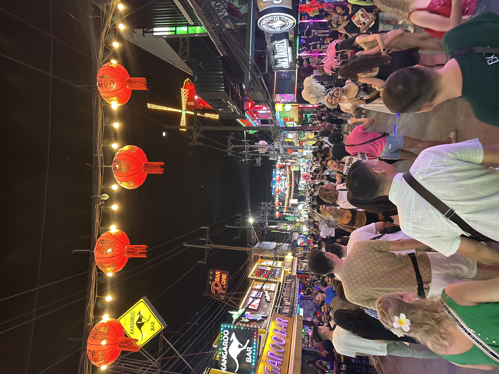 The image captures a crowded, illuminated street scene at night on Bangla Road in Patong, Phuket, Thailand. Overhead, red Chinese lanterns with gold characters and string lights are suspended across the thoroughfare, which is densely packed with pedestrians. The street is flanked by multi-story buildings housing numerous bars and entertainment venues, characterized by bright, colorful neon signage. Visible signs include "Kangaroo Bar," "P. BAR ONE," and "Jack Daniel's" branding, along with a distinct yellow neon windmill shape on the right side. A dense network of utility wires is visible against the dark sky.
A diverse crowd of people, mostly adults, fills the street, walking, standing, and interacting. Many are casually dressed. In the midground on the right, several individuals identified as performers are visible. One wears a white gown with a large white feathered headpiece, interacting with another in a dark sequined dress and a black feathered headpiece. Further right, another performer sports a pink feathered headpiece. Behind them, an elevated stage or platform with additional individuals, possibly dancers, is illuminated by pink and blue lights. In the foreground, the backs of several individuals are visible, including a person wearing a patterned beige shirt and white shorts, a woman with a white flower in her blonde hair wearing a green top, and a person in a white t-shirt. One person in a pink shirt has a "Take Photo" sign on their back. The scene is dynamic and bustling with activity.