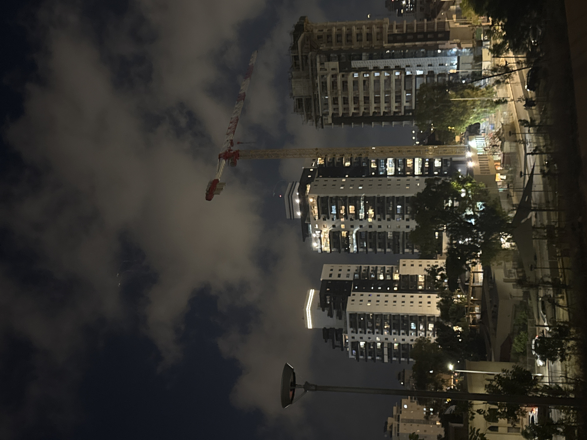 This is a nighttime photograph taken in Netanya, Israel, depicting a construction site adjacent to several tall residential buildings. A large construction crane with red and white markings dominates the upper portion of the frame, its boom extending horizontally. Several apartment buildings are visible, with lights on in many of their windows, indicating occupancy. One building is under active construction, showing exposed framework. In the foreground, a street lamp with a wide, shallow shade is positioned to the left of the frame. The sky is dark and cloudy, with some lighter patches suggesting moonlight filtering through. The scene appears to be a typical urban environment undergoing development.