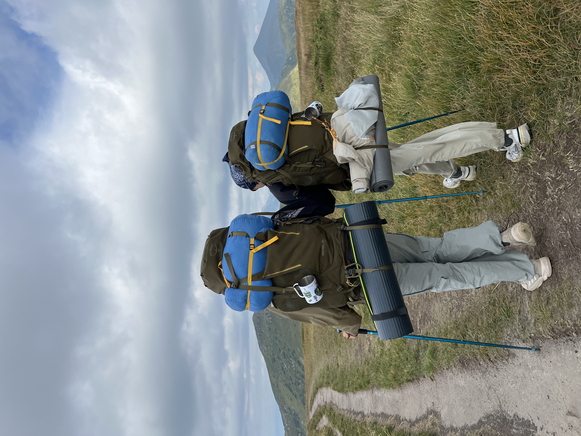 Two individuals, viewed from behind, are hiking on a dirt path in a mountainous landscape under a cloudy sky. The person on the left wears a green jacket, light grey cargo pants, and white sneakers, carrying a large olive green backpack with a blue sleeping bag attached and a rolled-up sleeping mat on the side. A white mug with a design is hanging from the backpack. They are using blue trekking poles. The person on the right wears a dark top, light beige cargo pants, and white sneakers, carrying a similar olive green backpack with a blue sleeping bag, a rolled-up sleeping mat, and folded clothing attached. They are also using blue trekking poles. The path winds through grassy hills, with distant, hazy mountains visible in the background. The scene is indicative of hiking or trekking in a rural, natural environment, likely in the Carpathian Mountains region, which includes the town of Vorokhta, Ukraine.