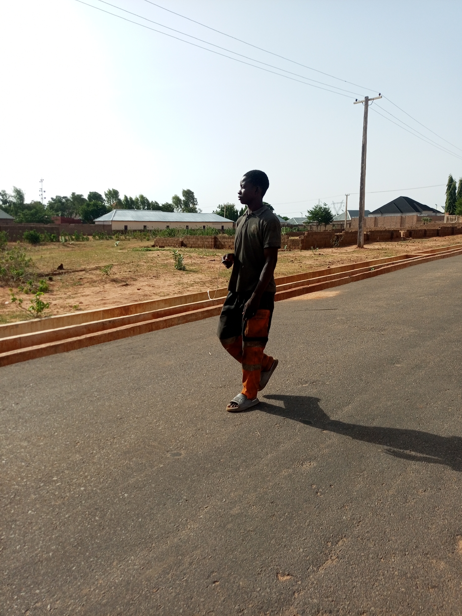 A young man walks on an asphalt road in Gombe, Nigeria. The road runs parallel to a low, red-brick curb and is lined with buildings, some with corrugated metal roofs. A utility pole stands near the side of the road. The sky is overcast.