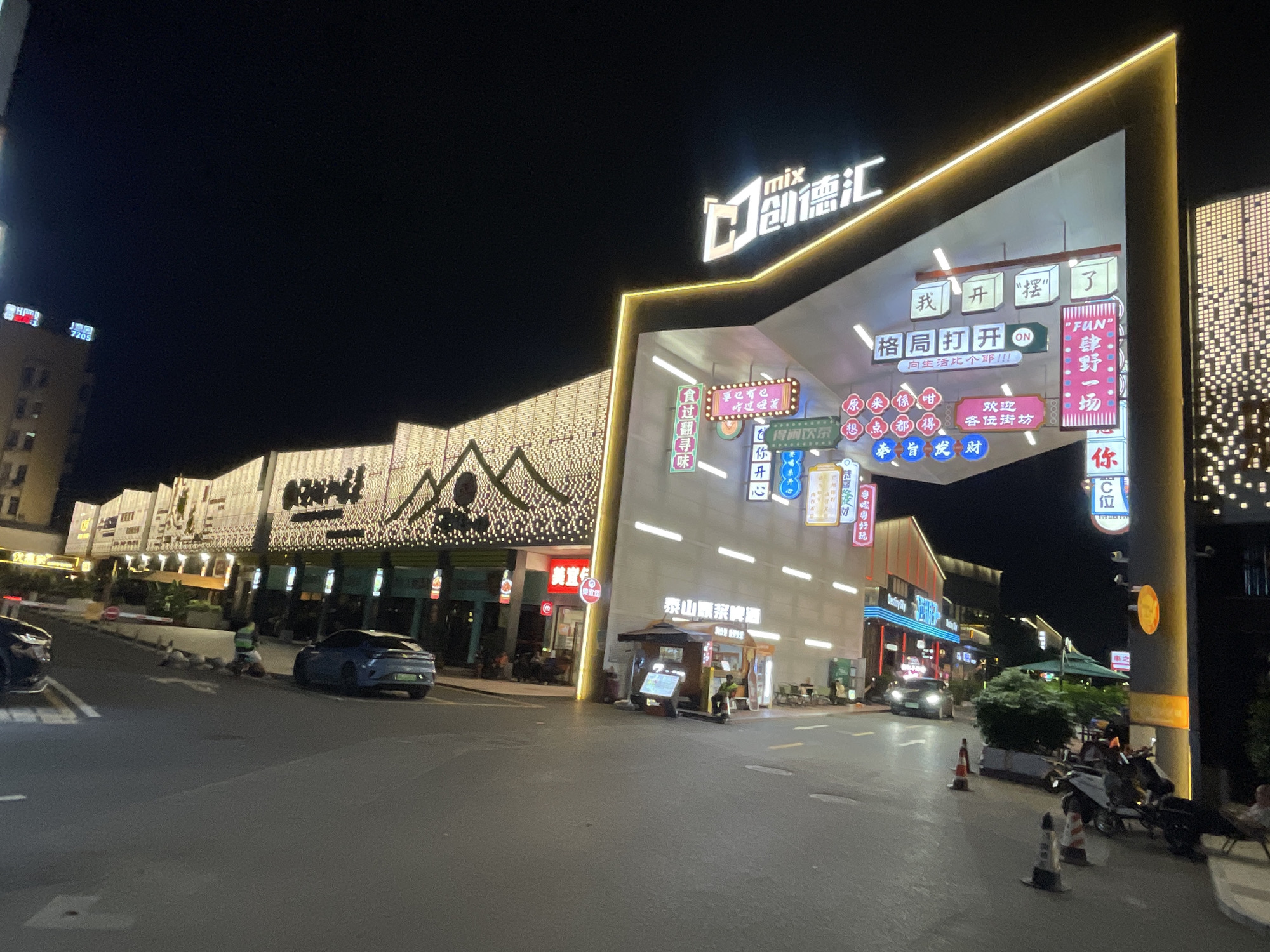 The image depicts a street scene at night in Panyu District, China. The foreground shows a paved road with several parked motorcycles and a blue sedan. Traffic cones are present on the road. A person in a high-visibility vest is visible near the blue car.
The background features a modern architectural structure with illuminated signage and decorative lights. This structure appears to be an entrance or a prominent building within a commercial area. Various businesses with neon signs are visible along the street, contributing to the vibrant nighttime atmosphere. Buildings are decorated with numerous small, bright lights, creating a festive ambiance. The sky is dark, indicating it is nighttime. The overall scene suggests a lively commercial or entertainment district.