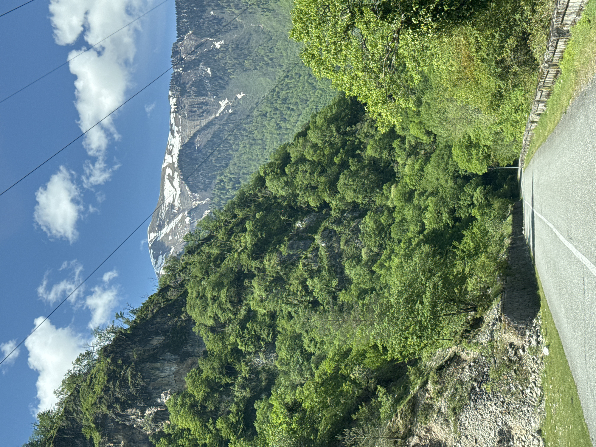 The image depicts a mountain scene, possibly taken from a moving vehicle. In the background, a snow-capped mountain rises against a blue sky dotted with cumulus clouds. Power lines traverse the upper left of the frame. The foreground is dominated by a steep, rocky cliff covered in dense green vegetation, primarily trees and shrubs. A paved road, marked with a white line, runs along the right edge of the image, adjacent to the vegetation. The location is likely Bzypta, Abkhazia, based on the prompt. There are no people visible. The interaction involves a road passing through the mountainous terrain. The environment is characterized by a combination of rocky cliffs, lush vegetation, and a mountainous backdrop under a partly cloudy sky.
