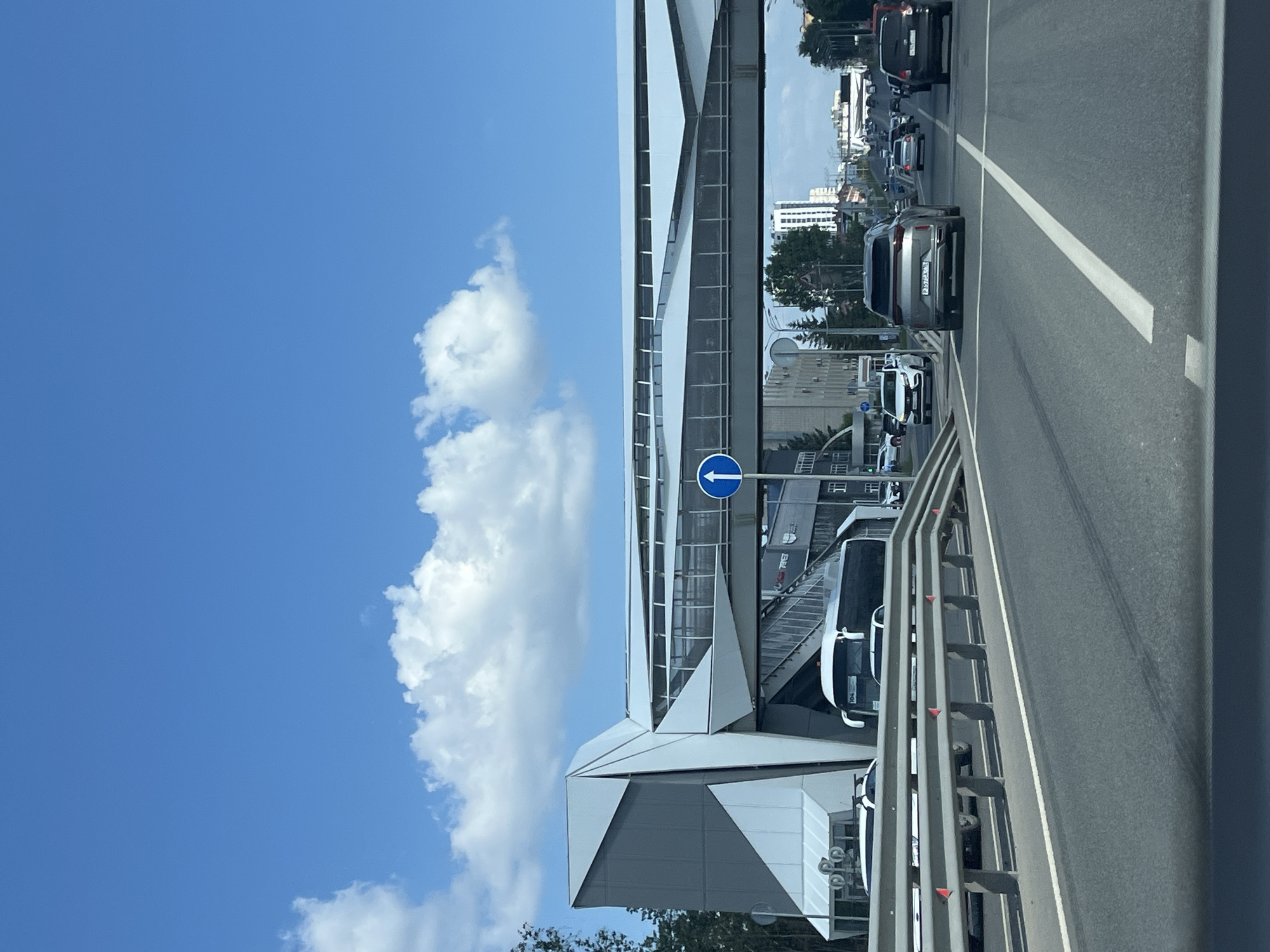 The image shows a street scene in Kazan, Russia, on a bright, sunny day with a blue sky and scattered white clouds. A modern, angular pedestrian bridge spans over a multi-lane road. Several cars and a bus are visible on the road. The pedestrian bridge has a grey and white geometric design. A blue circular sign with a white arrow pointing upwards is attached to the side of the bridge. The surrounding environment includes buildings, trees, and guardrails along the road. The perspective is from inside a vehicle traveling on the road.