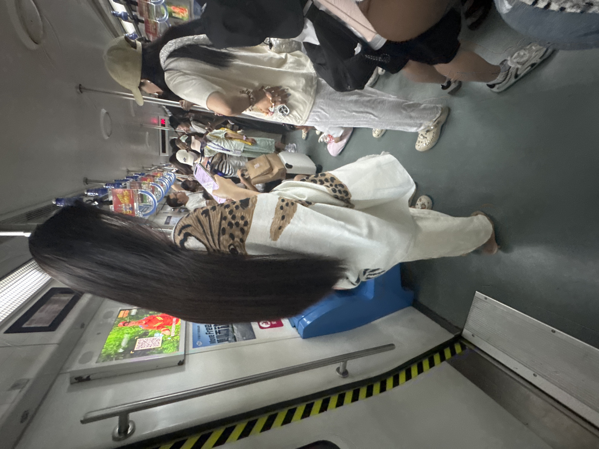The image shows the interior of a crowded subway car. In the foreground, a woman with very long, dark hair is standing, facing away from the camera. She is wearing a white outfit with a leopard print pattern on the top. She is holding a smartphone in her right hand. To her right, other passengers are visible, including a woman in a light-colored t-shirt and white pants, and another woman wearing a tan baseball cap. Some passengers are standing while others appear to be seated on the floor. The subway car has metal poles for support and advertisements displayed on the walls. The floor is a dark grey color, and there is a yellow and black striped hazard warning near the door. The lighting suggests it is daytime.