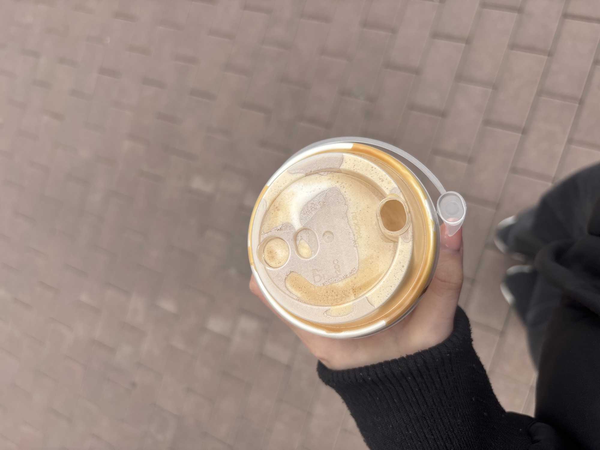 A person holds a cup of coffee with a plastic lid, while standing on a brick-paved surface that appears to be a sidewalk or pedestrian area. The person wears a black sweater and black leggings. The feet wear white and black tennis shoes. The scene may be taken in Saint Petersburg, Russia.