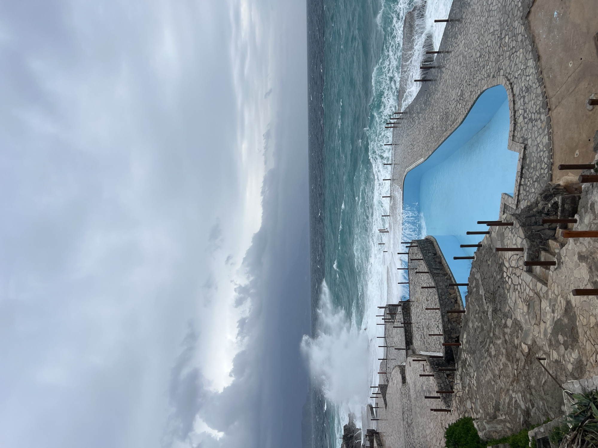 The image displays a turbulent coastal scene in Krimovica, Montenegro, under a heavily overcast sky dominated by dark gray clouds. A light blue, irregularly shaped swimming pool, appearing calm, is centrally positioned in the foreground and mid-ground, integrated into a multi-tiered stone-paved coastline. The adjacent sea exhibits turbulent, greenish-blue water with significant wave activity. A large wave is actively breaking on the left side of the frame, generating substantial white spray against the rocky formations and lower stone platforms. Smaller waves are observed washing over the stone walkways immediately bordering the pool, indicating powerful sea conditions. These walkways and terraces are constructed from irregular gray stone and appear wet from the ocean spray. Numerous short, dark brown posts delineate the edges of the paved areas and surround the pool. Sparse green coastal vegetation is present in the lower left section. No human or animal figures are present in the scene.