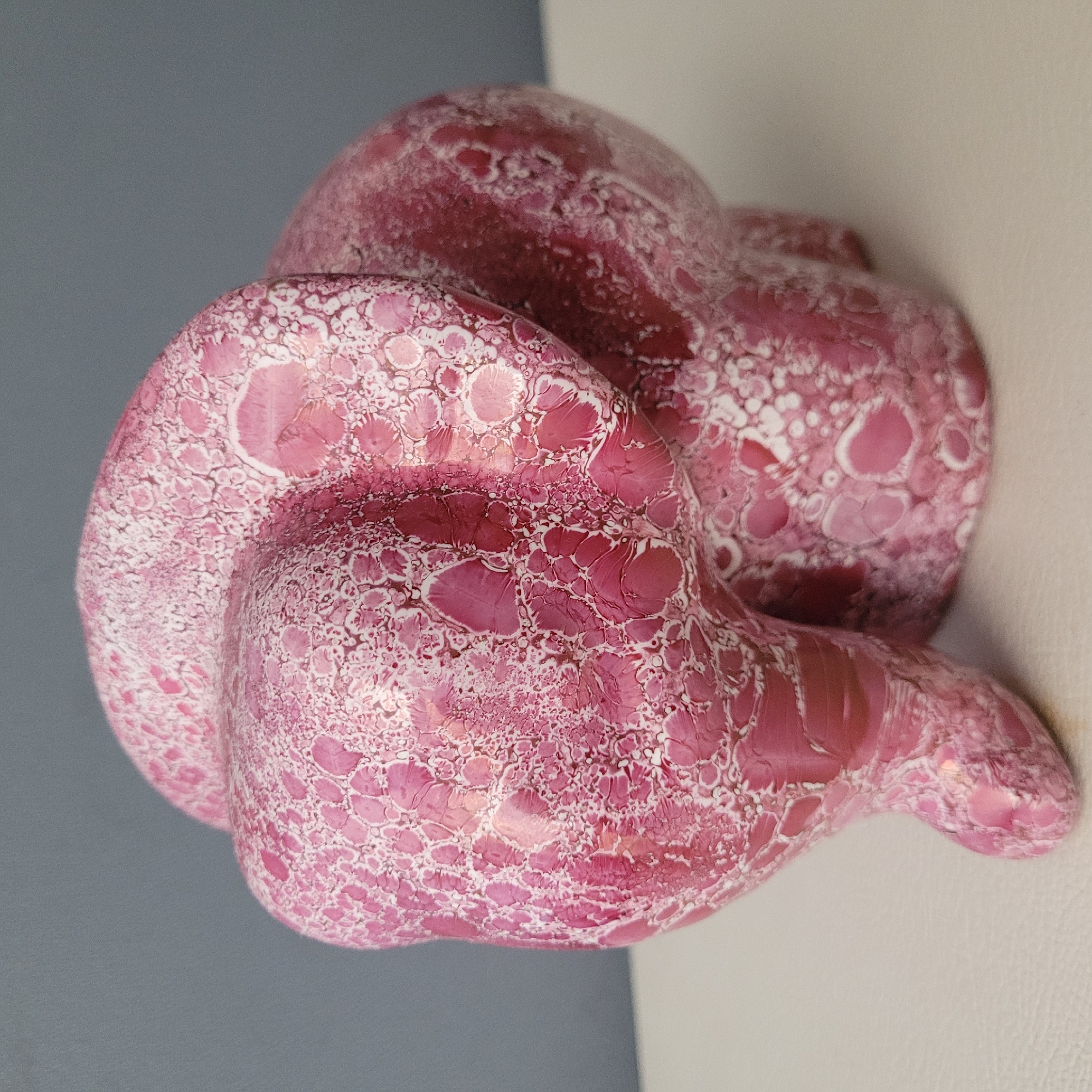 The image shows a close-up of a decorative pink elephant figurine. The figurine has a textured surface with a marbled or bubbly pattern in shades of pink and white. The background consists of a plain gray wall and a white surface the figurine is resting on. There are no people or animals present in the image. The location of the image in Jūrmala, Latvia cannot be determined from the provided visual information.