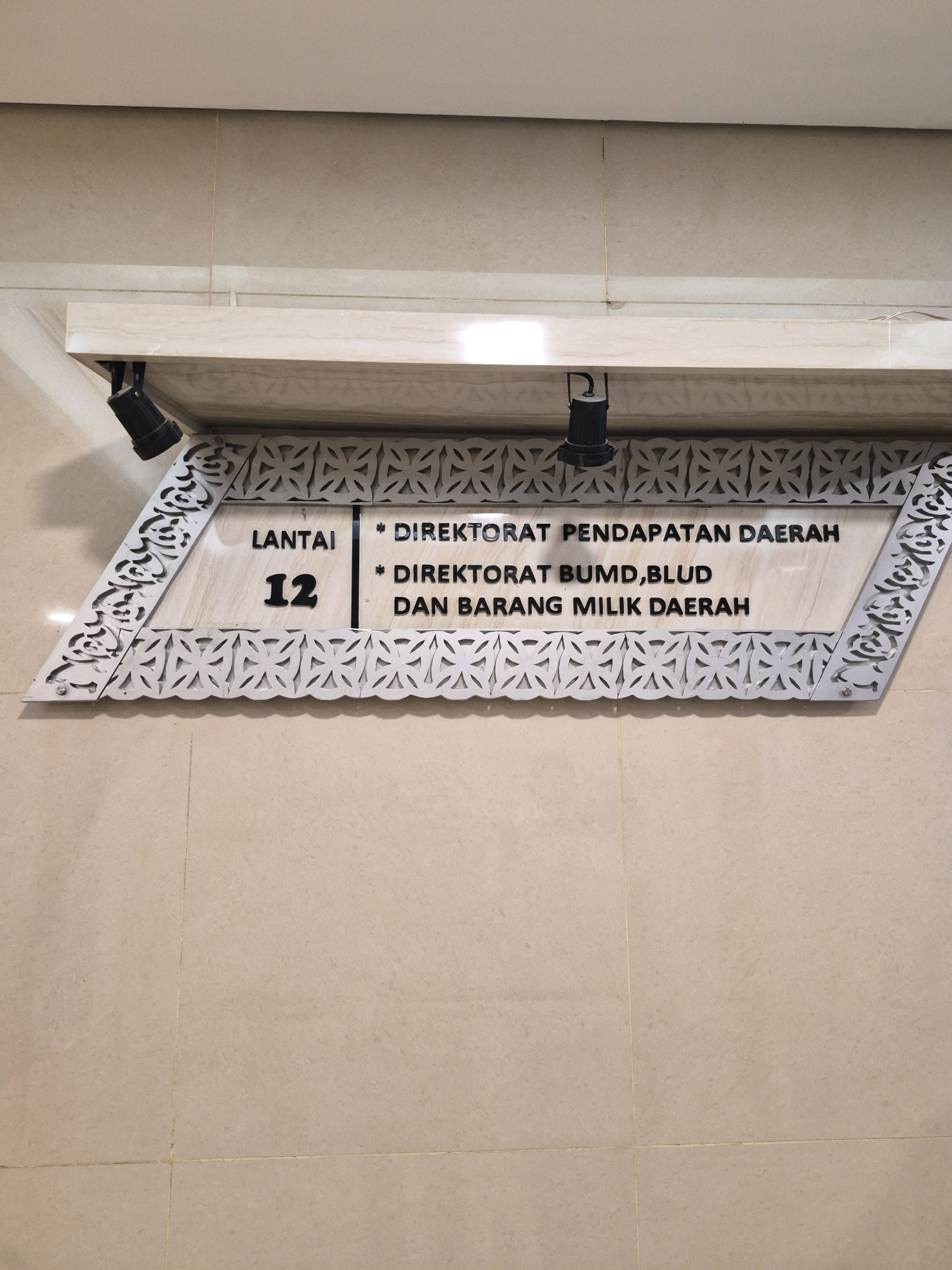 The image shows a sign on a marble-like wall, indicating "LANTAI 12" (Floor 12) and "* DIREKTORAT PENDAPATAN DAERAH" (* Regional Revenue Office) and "* DIREKTORAT BUMD, BLUD DAN BARANG MILIK DAERAH" (* Directorate of BUMD, BLUD, and Regional Owned Assets). The sign has decorative carved elements on its sides and is illuminated by two black spotlights mounted on a shelf above. The wall is tiled with light beige tiles. This sign is likely located in a government building in Central Jakarta, Indonesia, given the Indonesian language and the nature of the office names, which are typical of regional government structures. There are no people visible in the image.