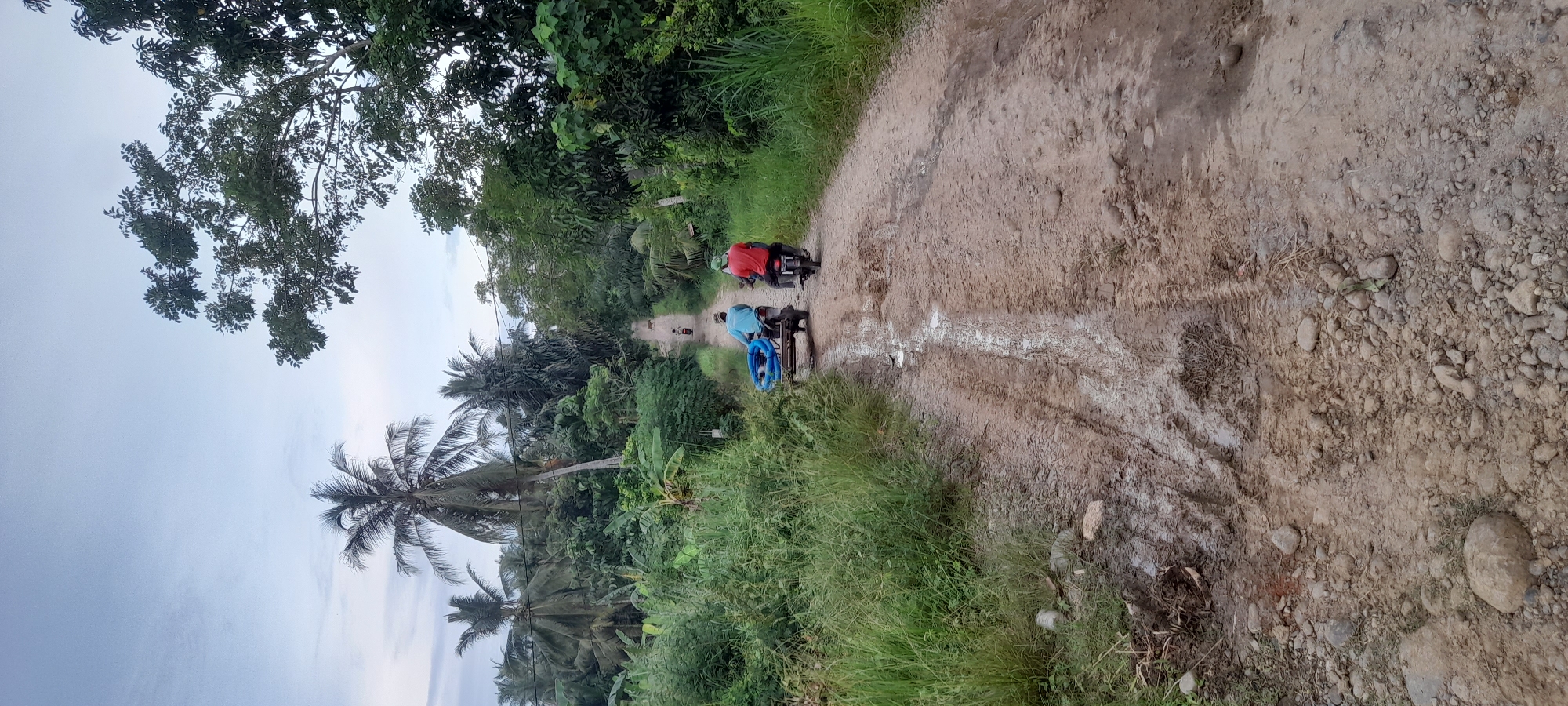 The image depicts a dirt road in Uteunbunta, Indonesia, lined with dense tropical vegetation. Two motorcycles are traveling away from the viewer along the road. The first rider wears a red shirt, and the second wears a blue shirt with blue coiled material strapped to the back. The road appears muddy in places, with visible tire tracks. The sky is overcast.