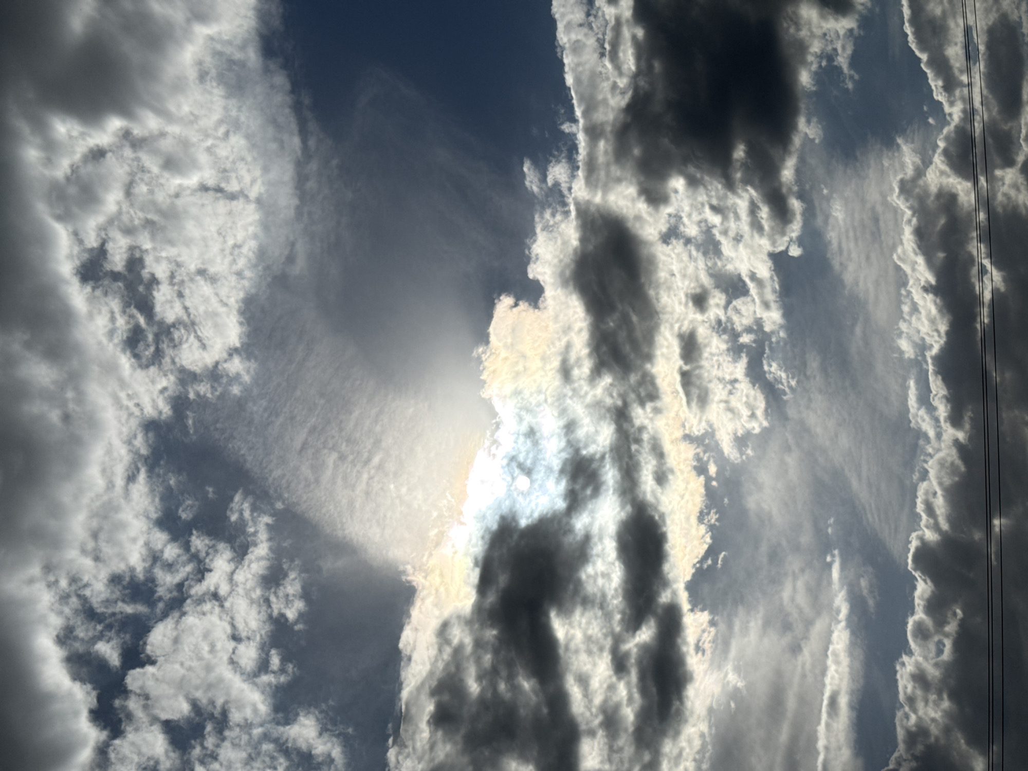 The image shows a daytime sky in Chaikovsky, Russia, filled with various cloud formations. The clouds range from thin, wispy cirrus clouds to denser cumuliform clouds, with the sun partially obscured by them. The sunlight breaks through in some areas, creating a bright, diffused glow. The colors range from dark grays and whites to the blue of the sky visible in gaps between the clouds. Power lines are visible on the right side of the image.