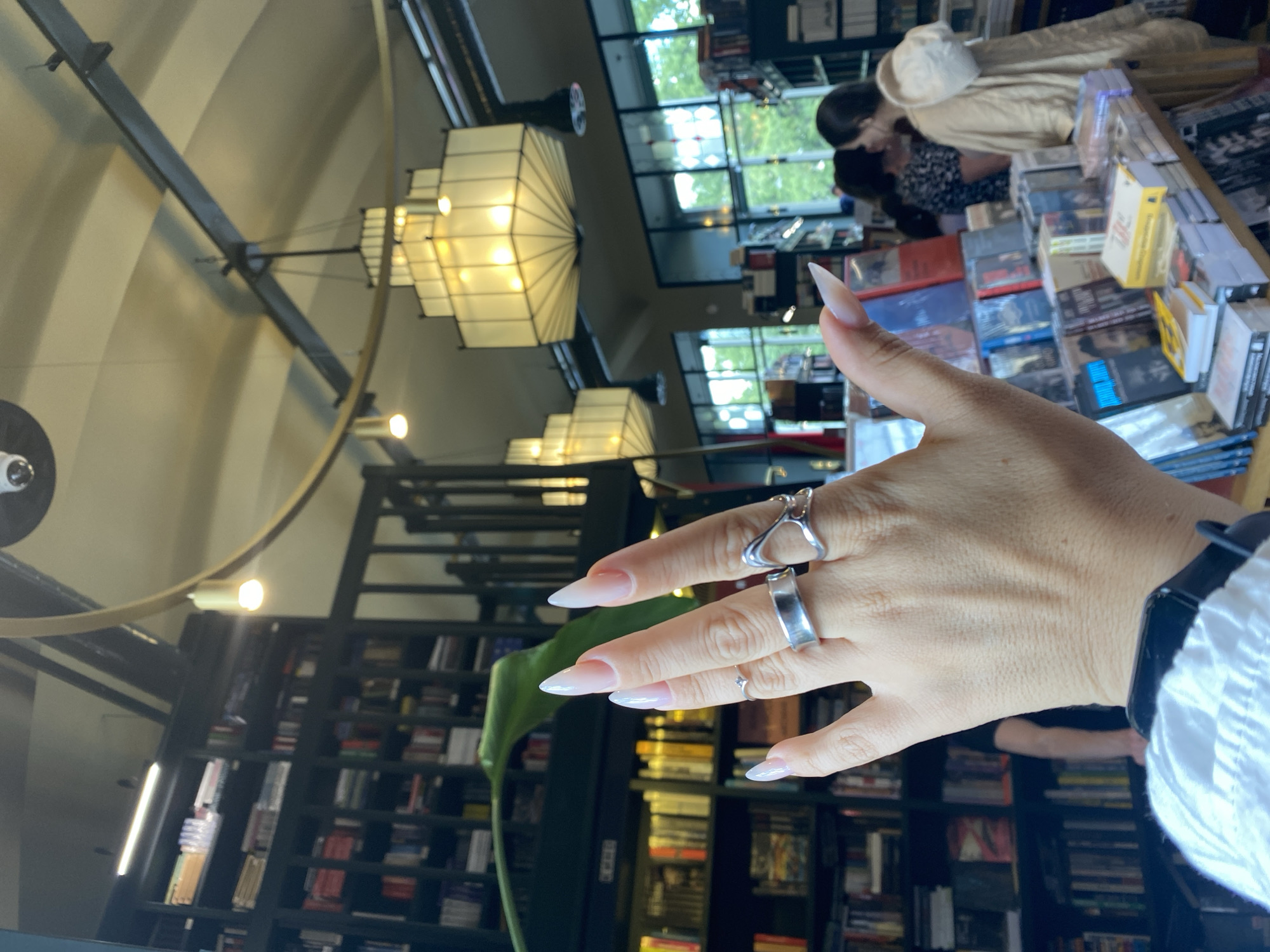 The image depicts an interior scene within what appears to be a bookstore. In the foreground, a hand with long, pointed, light pink-toned nails is raised, displaying several silver rings. The hand is positioned in front of a lush green plant leaf.
The background reveals multiple levels of dark wooden bookshelves densely packed with books. Several hanging light fixtures, one a prominent multi-tiered rectangular lantern with warm illumination, and another smaller cylindrical light, are suspended from the ceiling, which features exposed beams and curved surfaces.
Through large windows in the middle ground, glimpses of greenery and natural light suggest an outdoor environment beyond the building. In the further background, several individuals are visible browsing, including two women in conversation near a display of books. The overall ambiance suggests a well-lit, possibly academic or cultural establishment. The image does not contain information about the specific location within Saint Petersburg, Russia.