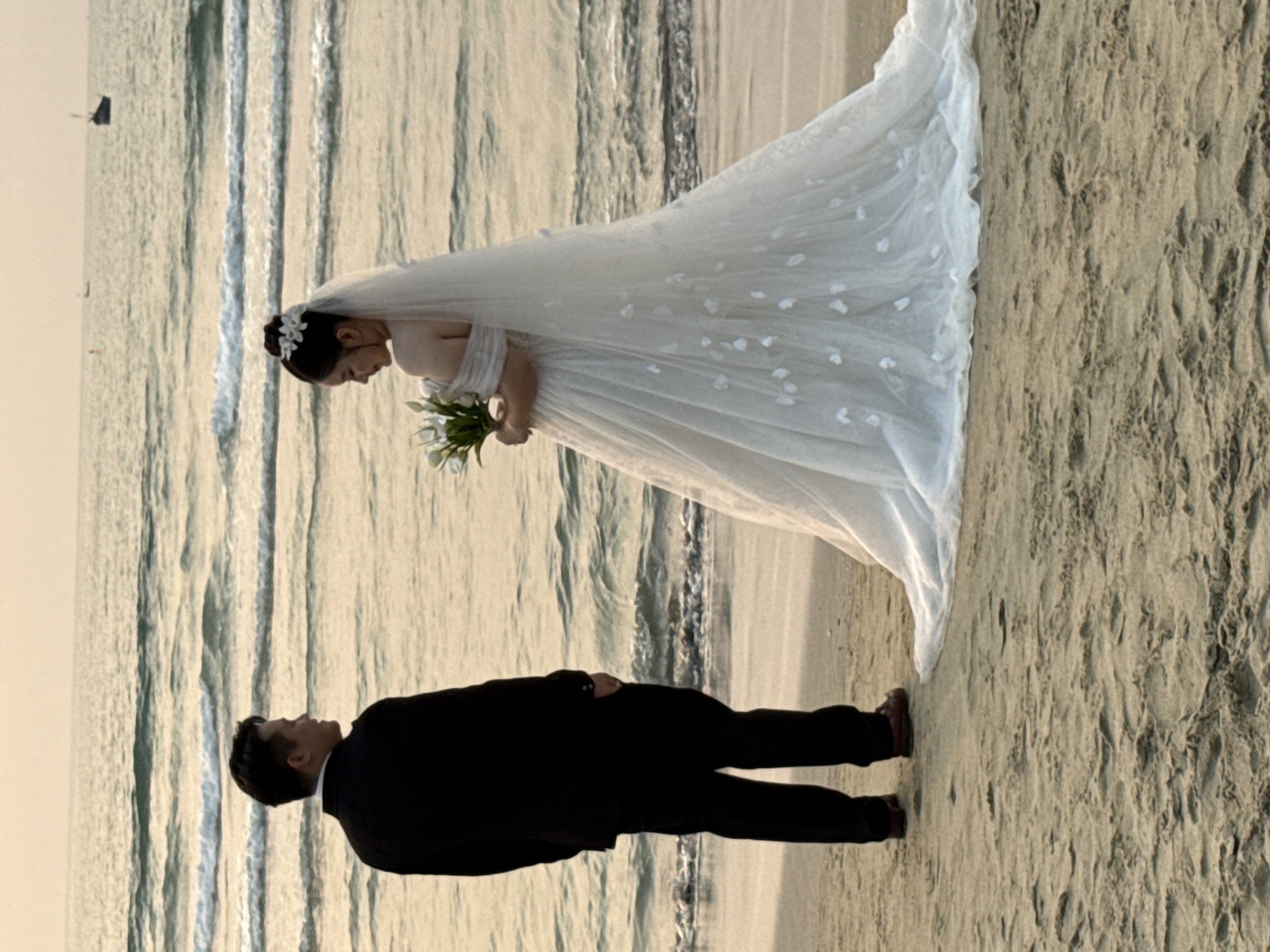 A wedding photo shows a bride and groom on a beach. The bride is wearing a white wedding dress with a veil and holding a bouquet of white flowers. The groom is wearing a black suit. They are standing close together on the sand. The background includes the ocean with small waves and possibly a boat in the distance. Based on the coastal environment, it is plausible the location is a beach in Đà Nẵng, Vietnam.