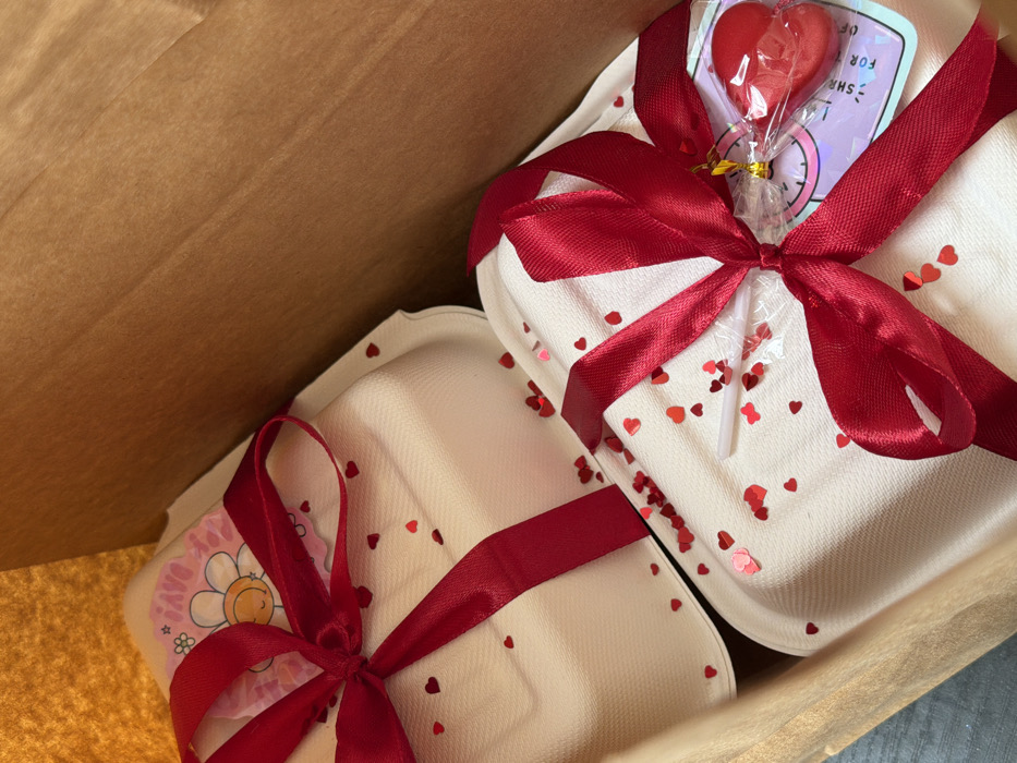 Two off-white, textured rectangular containers, possibly compostable takeout boxes, are depicted inside a brown paper bag. Each container is adorned with a red satin ribbon tied into a bow. Scattered around both containers are numerous small, red, heart-shaped confetti pieces. The upper container features a pink, scalloped-edged sticker displaying a cartoon daisy and partially visible text. The lower container has a clear cellophane-wrapped red heart-shaped lollipop attached to its ribbon, along with a square, iridescent tag. The interior of the brown paper bag is visible, with a darker surface beneath the bag visible in the lower left corner. No individuals are present. The image itself contains no visual information to confirm a specific location within the city of Tel Aviv-Yafo, Israel.