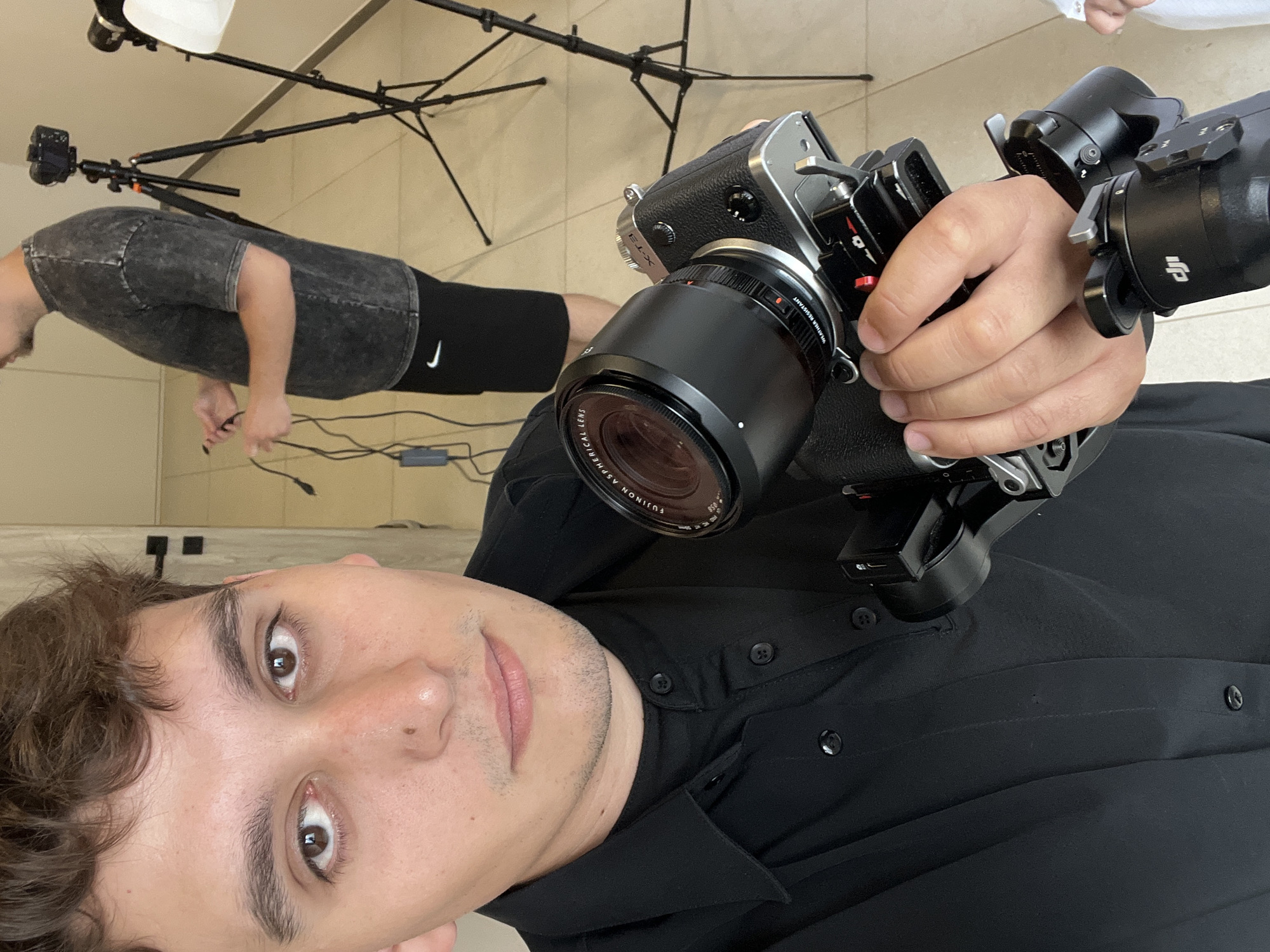 An indoor, vertically oriented image captures a young man holding a professional camera setup, likely in a studio or production environment in Zelenodol'sk, Russia.
In the foreground, a man with short brown hair, dark brown eyes, and light stubble looks directly at the viewer with a neutral expression. He is wearing a black button-up shirt. His right hand grips a silver and black Fujifilm X-T3 camera, which is fitted with a black FUJINON ASPHERICAL LENS. The lens barrel shows "WR" (Weather Resistant). The camera is integrated into a larger black rig or gimbal system, prominently displaying the "DJI" logo on its components.
In the background, another person is partially visible, wearing a dark grey t-shirt with a faded, acid-wash appearance and black athletic shorts or leggings featuring a white "Nike" swoosh logo. This individual is positioned near the floor, seemingly engaged with cables.
The setting comprises light-colored tiled flooring and plain walls. Several black light stands or tripods are visible, one supporting a white reflector or softbox, indicative of a photography or videography studio setup. Cables are strewn across the floor. The overall scene suggests a behind-the-scenes moment or a self-portrait taken during a production shoot.