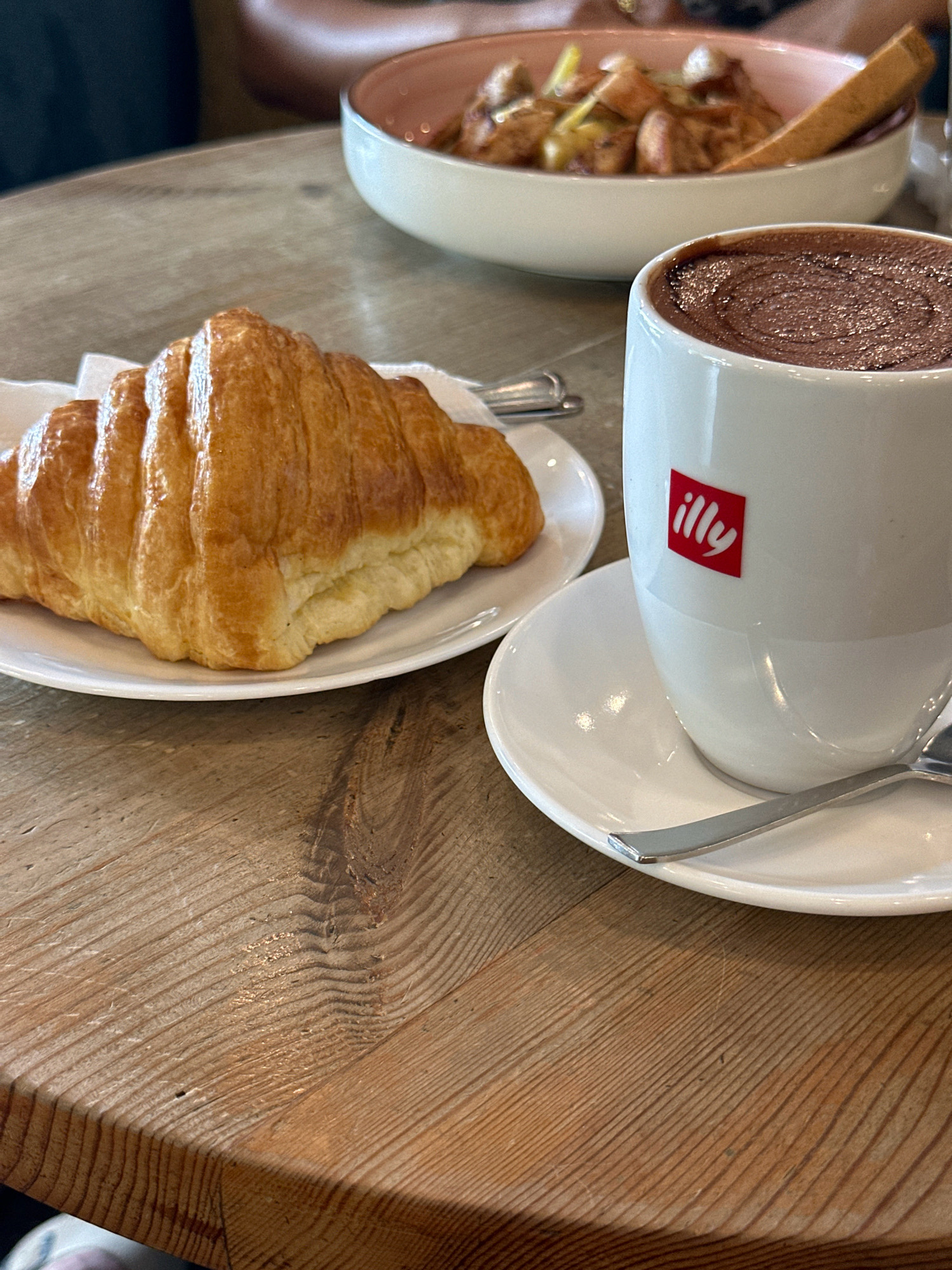 In Kathmandu Metropolitan City, Nepal, a close-up shot captures a breakfast scene on a wooden table. A golden-brown croissant rests on a white plate, positioned to the left of a white Illy coffee cup filled with dark chocolate. The cup, with its distinctive red Illy logo, sits on a matching saucer, accompanied by a silver spoon. In the blurred background, a pink-rimmed bowl contains a dish of food with what appears to be bread and vegetables. The wooden surface of the table displays a prominent grain pattern, suggesting it is a well-used piece of furniture.