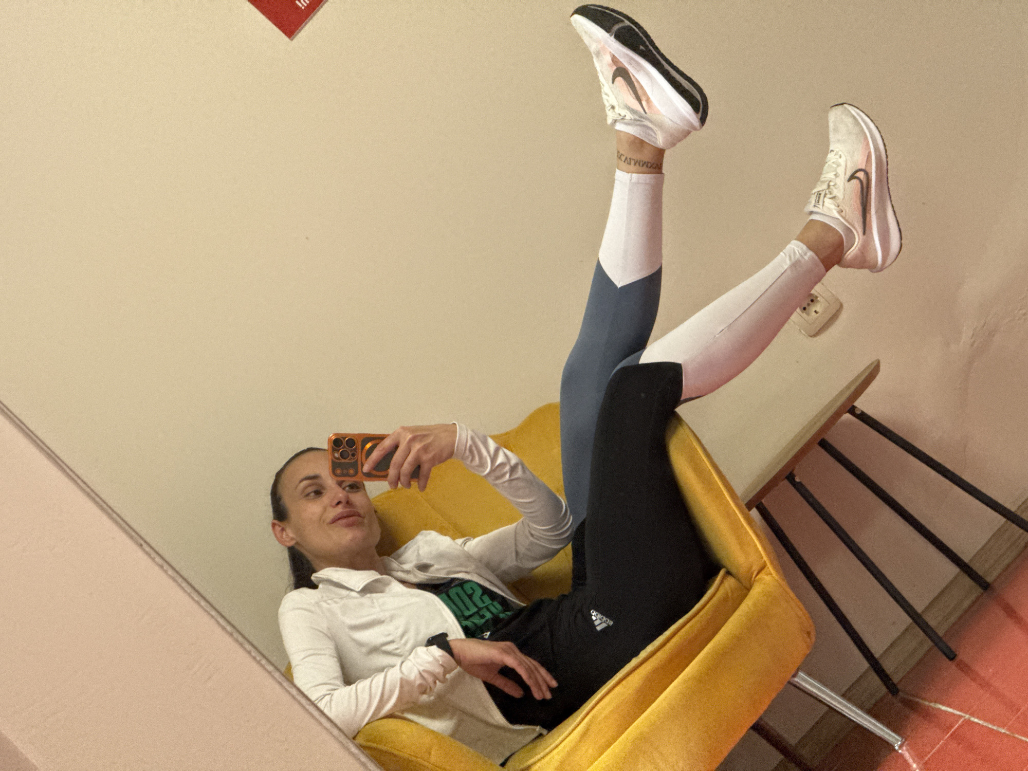 A woman is reclined in a yellow upholstered chair with her legs raised vertically. She is holding a phone and appears to be taking a selfie in a mirror. The woman is wearing running shoes, leggings, a t-shirt, and a button-down jacket. One leg is in blue and white leggings, and the other is in black Adidas leggings. A tattoo is visible on one ankle. The background includes a light yellow wall, an electrical outlet, a metal-legged table, and a patterned floor, indicating the scene takes place indoors. Pleven, Bulgaria cannot be confirmed as the location based on the available visual information.