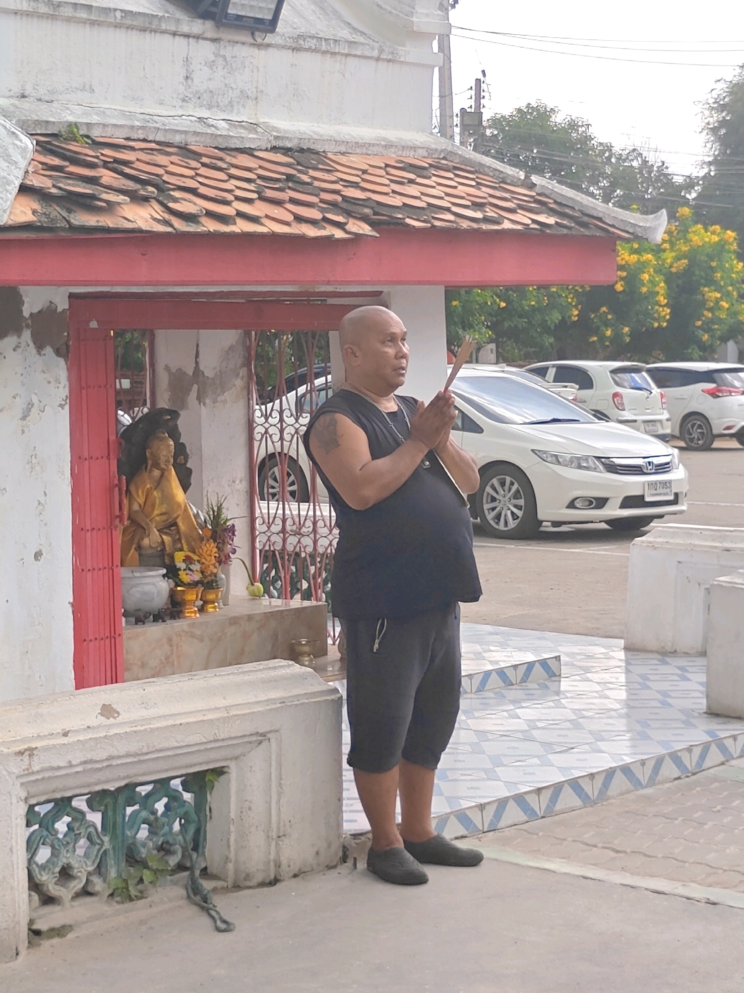 A bald man wearing a black tank top and dark shorts stands in front of a small shrine. He is holding incense sticks in his hands, which are pressed together in a prayer gesture. The shrine contains a golden Buddha statue, flowers, and other offerings. The man is standing on a tiled walkway, with a concrete structure in the foreground. In the background, there are white cars parked. The architecture suggests a location in Thailand, likely Phra Nakhon Si Ayutthaya City Municipality, known for its historical temples and religious sites. The overall scene depicts a moment of religious devotion.