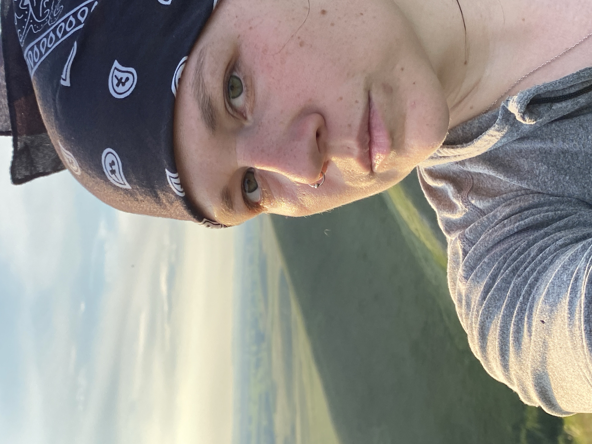 A medium close-up selfie shows a person with a black bandana with white paisley print on their head. The person has green eyes, a nose ring, and fair skin with freckles. They are wearing a gray hoodie and a silver necklace. The background is a blurred landscape of green fields under a hazy sky, with the shadow of a hill falling across the field. The scene appears to be outdoors, possibly in the city of Novogafarovo, Russia.