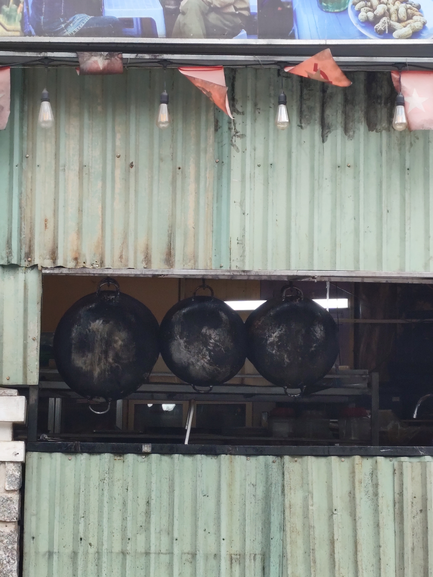 The image depicts a stall or eatery in Da Lat, Vietnam, featuring a corrugated metal facade painted a pale green color, showing signs of age and weathering. Above a horizontal opening, a string of bare Edison-style light bulbs hangs, interspersed with small, triangular red and white flags, possibly Vietnamese national flags. Three large, black, spherical woks are suspended by metal handles from a metal bar within the opening. These woks appear well-used, with visible dark residue. The background visible through the opening is dimly lit, revealing shelves and indistinct objects, suggesting an indoor preparation area. To the left of the woks, a stack of white and gray block-like structures are visible, possibly part of the stall's construction or additional shelving. The overall impression is of a functional, perhaps slightly rustic, food vendor setting.