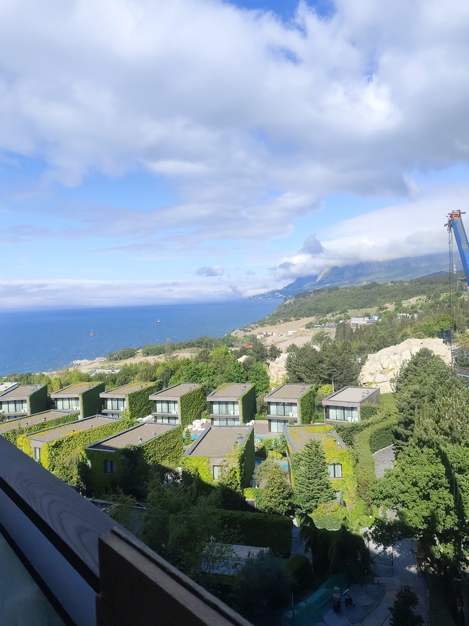 The image shows an aerial view of a coastline with modern, cube-shaped buildings covered in greenery. The buildings are situated on a hill overlooking the sea. In the background, there are mountains partially covered in clouds. The location is in the city of Симеизский поселковый совет, Ukraine. A blue construction crane is visible on the right side of the image. In the foreground, a dark-colored railing is visible. There is a clear sky with scattered clouds. There are a few boats in the sea.