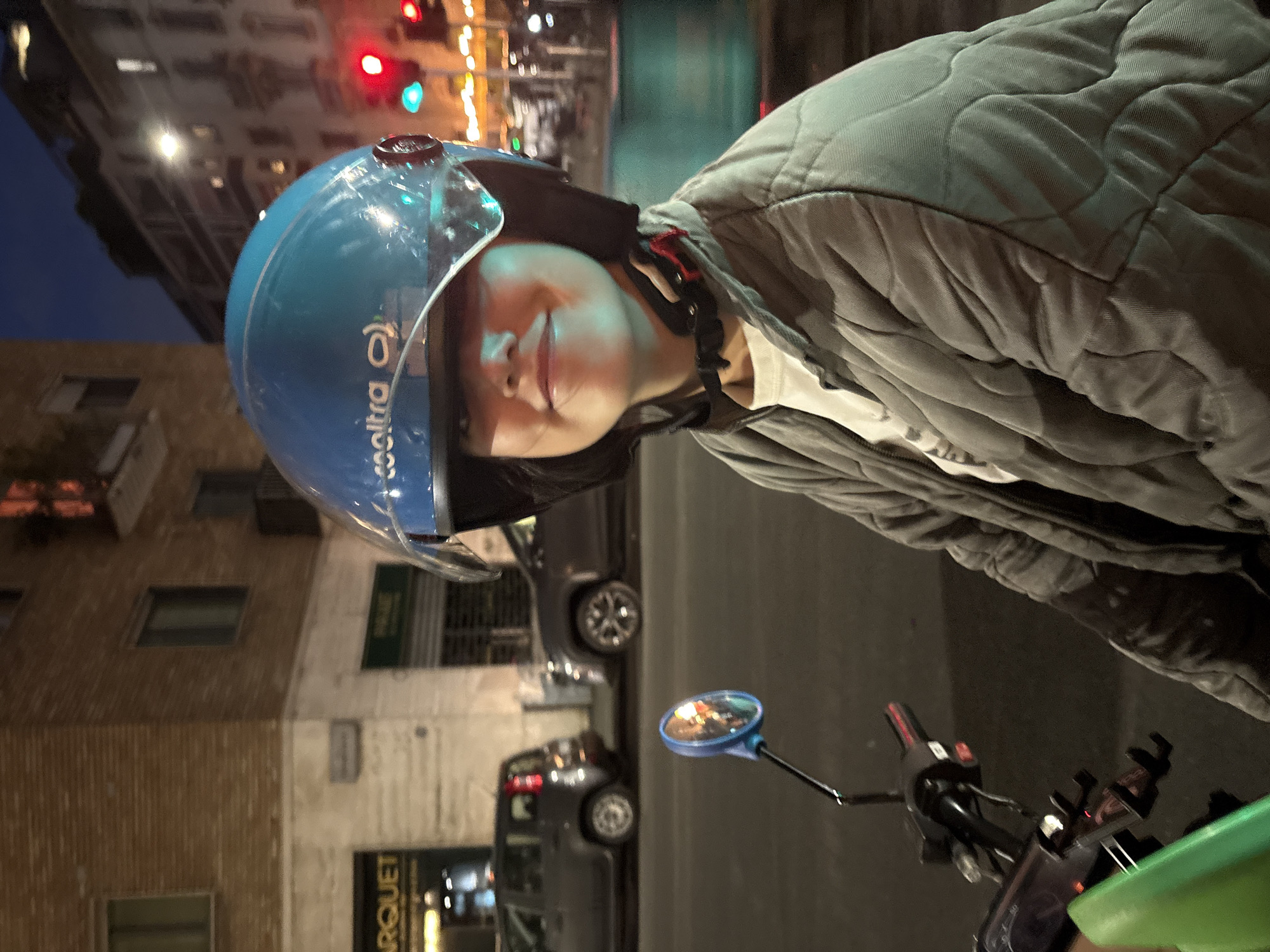 This nighttime or dusk photograph captures a person from the chest up, wearing a light blue helmet with a clear visor and a grey, quilted jacket. The person has a calm expression with a faint smile, looking slightly off-camera. The helmet features the word "cooltra" printed in white letters on its side.
The subject appears to be on a scooter or moped, evidenced by a blue-framed rearview mirror and black handlebars visible in the foreground. The background is an urban street scene with tall buildings lining the street, some with illuminated windows. Red and green traffic lights are visible further down the street, suggesting an active thoroughfare. Artificial lighting from streetlamps and building interiors casts a glow over the scene. Several cars are parked or moving in the background. On one of the buildings, a shop sign can be partially seen with the text "ARQUET". The overall ambiance suggests a typical evening in a city environment, such as Milan, Italy.