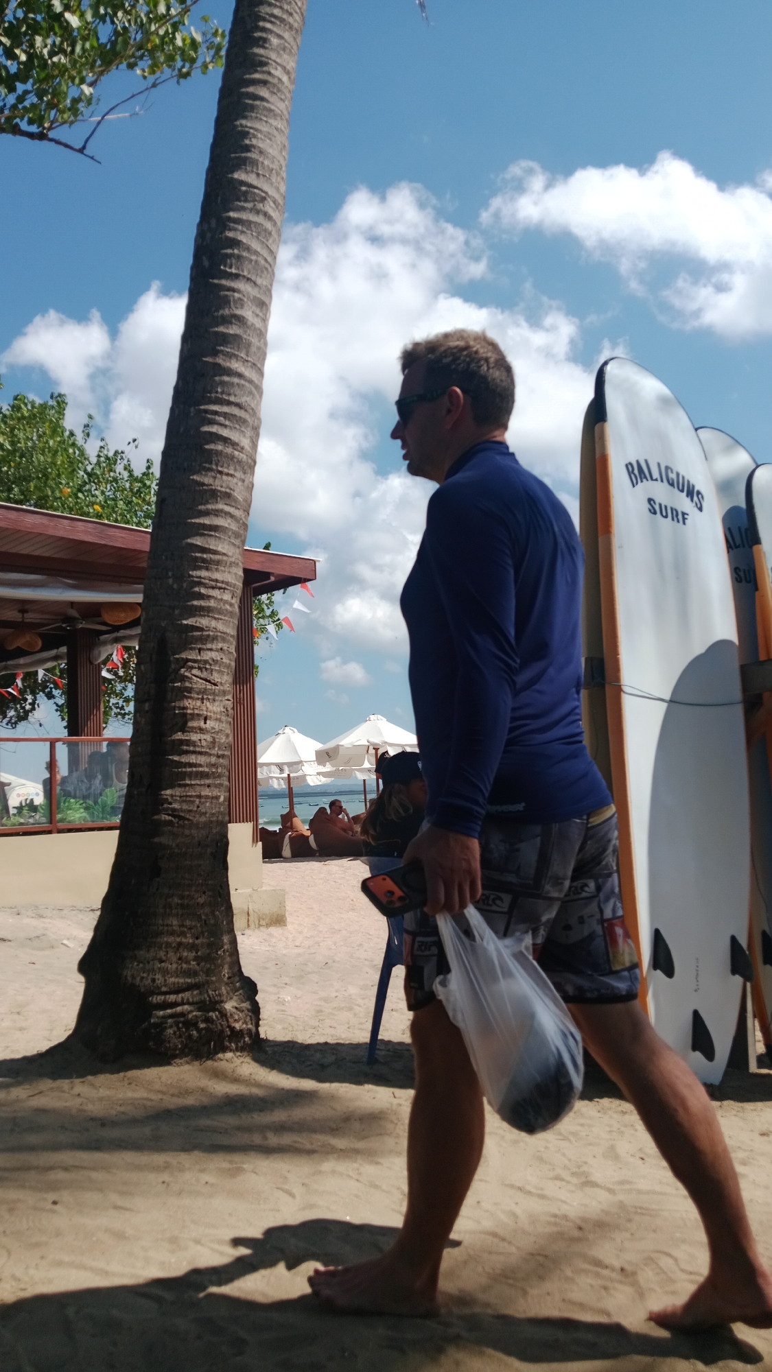 In Kuta, Indonesia, a man wearing a blue long-sleeved shirt and patterned shorts walks on a sandy beach. He is holding a white plastic bag in his right hand, which contains a dark object. The man has sunglasses on and is walking away from the camera. Behind him, several surfboards are propped up, one with the text "BALIGUNS SURF". Further in the background, there are beach umbrellas, lounge chairs, and people relaxing on the sand. A large palm tree trunk is on the left side of the image. The sky is blue with scattered clouds.