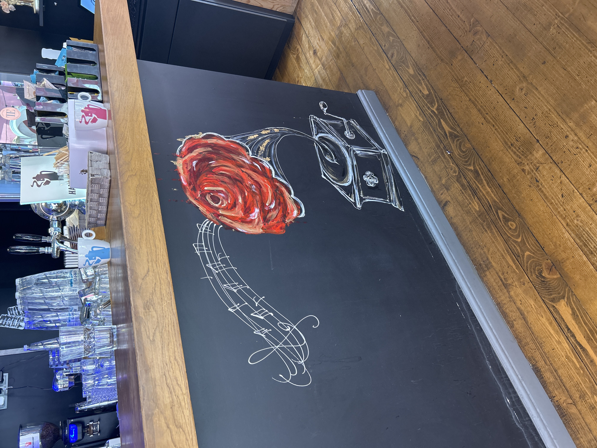 An indoor scene, likely a bar or café counter, is depicted from a slightly elevated angle. A warm-toned wooden counter runs diagonally from the lower left to the upper right.
On the black, chalkboard-like surface of the counter, a prominent artistic drawing features a vibrant red rose with golden accents. Flowing from the rose is a white, chalk-style rendering of a gramophone horn, which connects to an outlined gramophone speaker box. Below this, a musical stave with various white musical notes and clefs is drawn, suggesting a theme of music or melody.
To the left of the wooden counter, in the bar area, several items are visible. These include multiple shiny beer taps, an array of clear drinking glasses, some illuminated with a subtle blue light, and a white ceramic mug adorned with a blue, stylized figure. Various holders contain bar essentials like straws or condiments, with the partial text "HA" visible on one paper insert. A coffee grinder is also present further back.
To the right of the counter, a rustic wooden floor with distinct planks and grain is visible. A thin grey trim separates the black counter surface from the floor. No individuals are present in the image. On one of the clear glasses, a faint Cyrillic word, possibly "БЯР" or "БЯРХ", can be seen. The lighting is bright, indicating an active establishment or a well-lit interior.